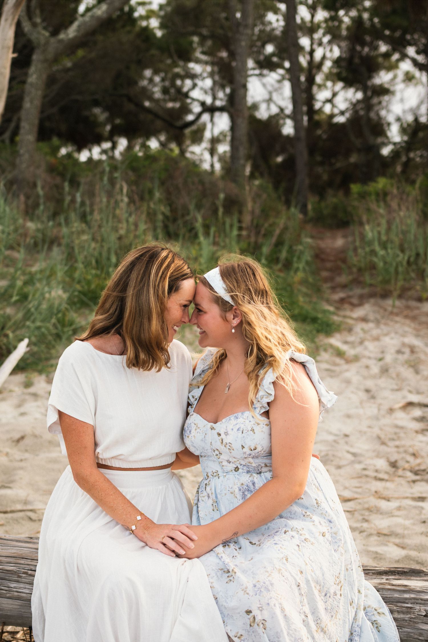 Two women in white and floral dresses sit together on a beach, foreheads touching in an intimate moment.