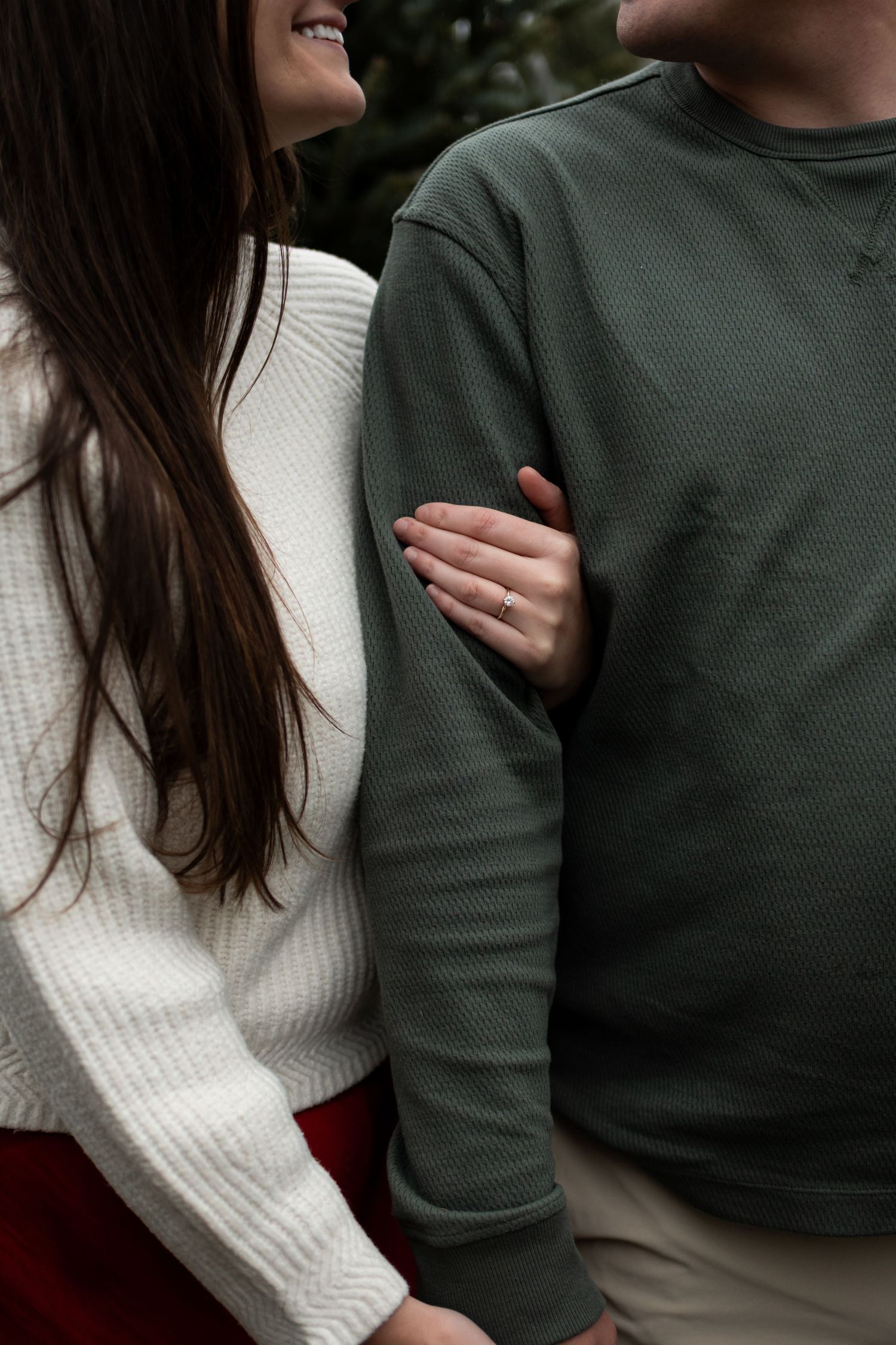 Couple in white knit and green thermal sweaters embracing outdoors, woman wearing engagement ring.