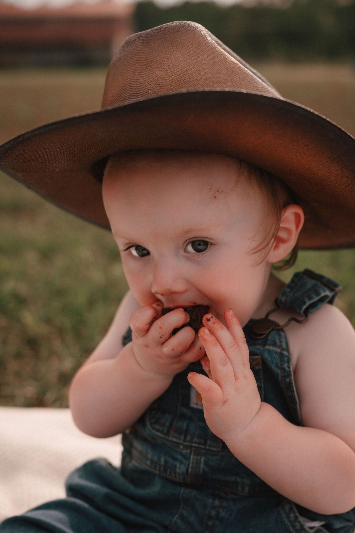 A cute baby in denim overalls and a brown cowboy hat eating a snack outdoors in a grassy field.