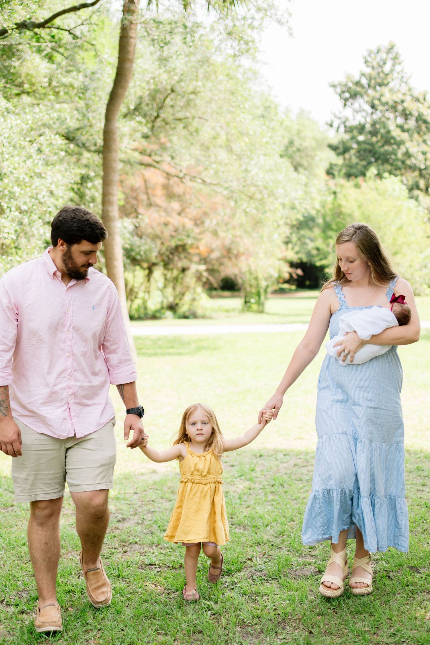 Family of four walking in a green park, parents holding toddler girl's hands, mother carrying newborn baby.