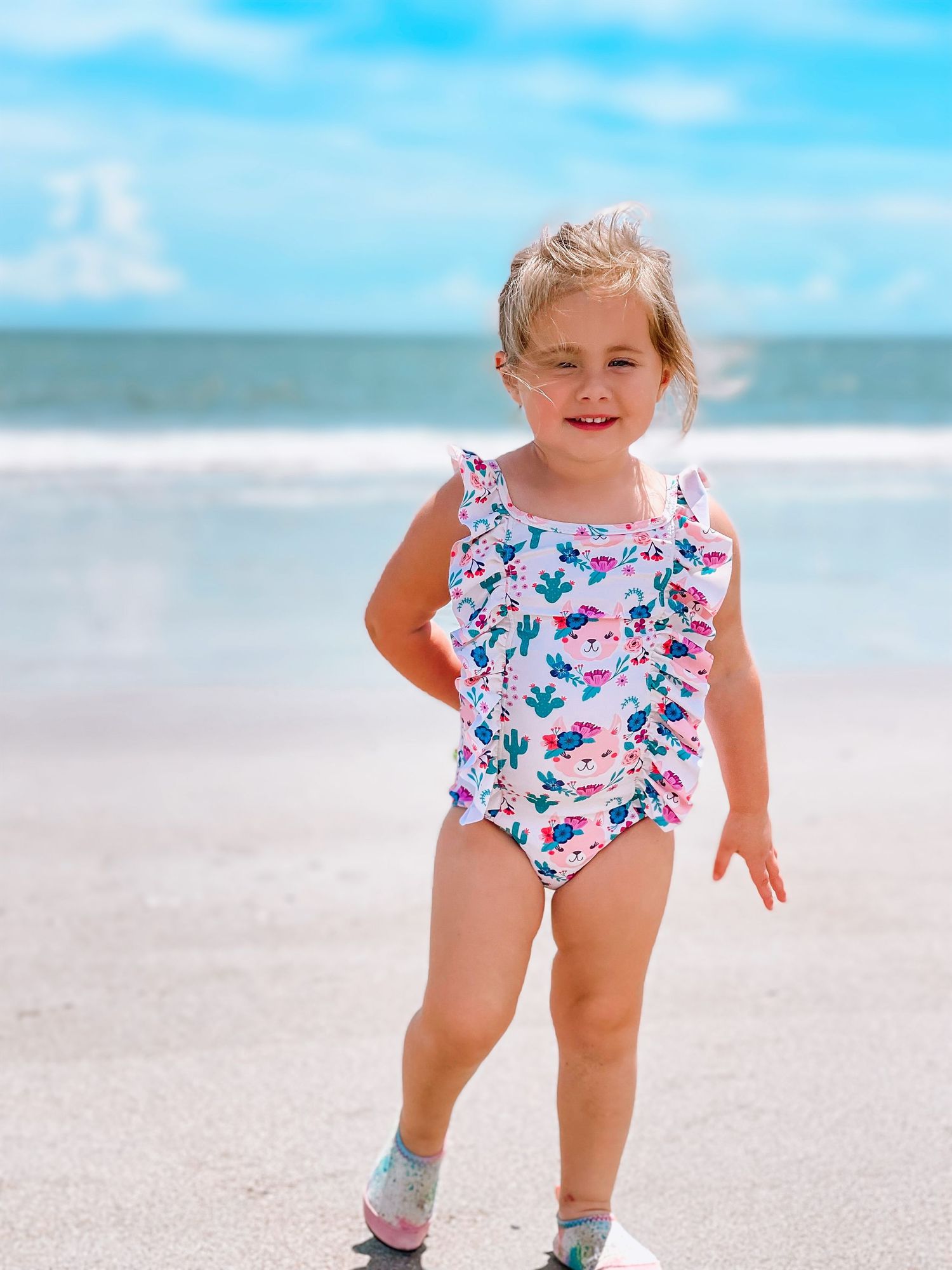 Adorable blonde toddler in pink floral ruffle swimsuit walking on sunny beach shoreline.