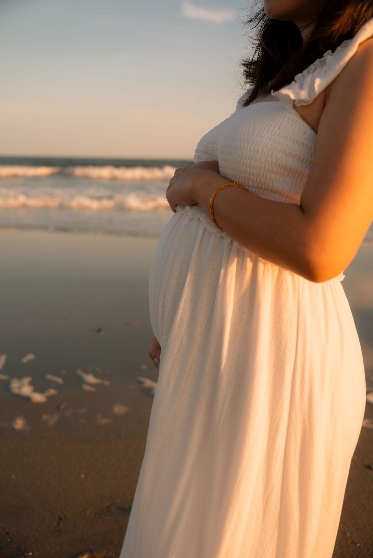 Pregnant woman in white dress cradling baby bump on beach at golden hour sunset.