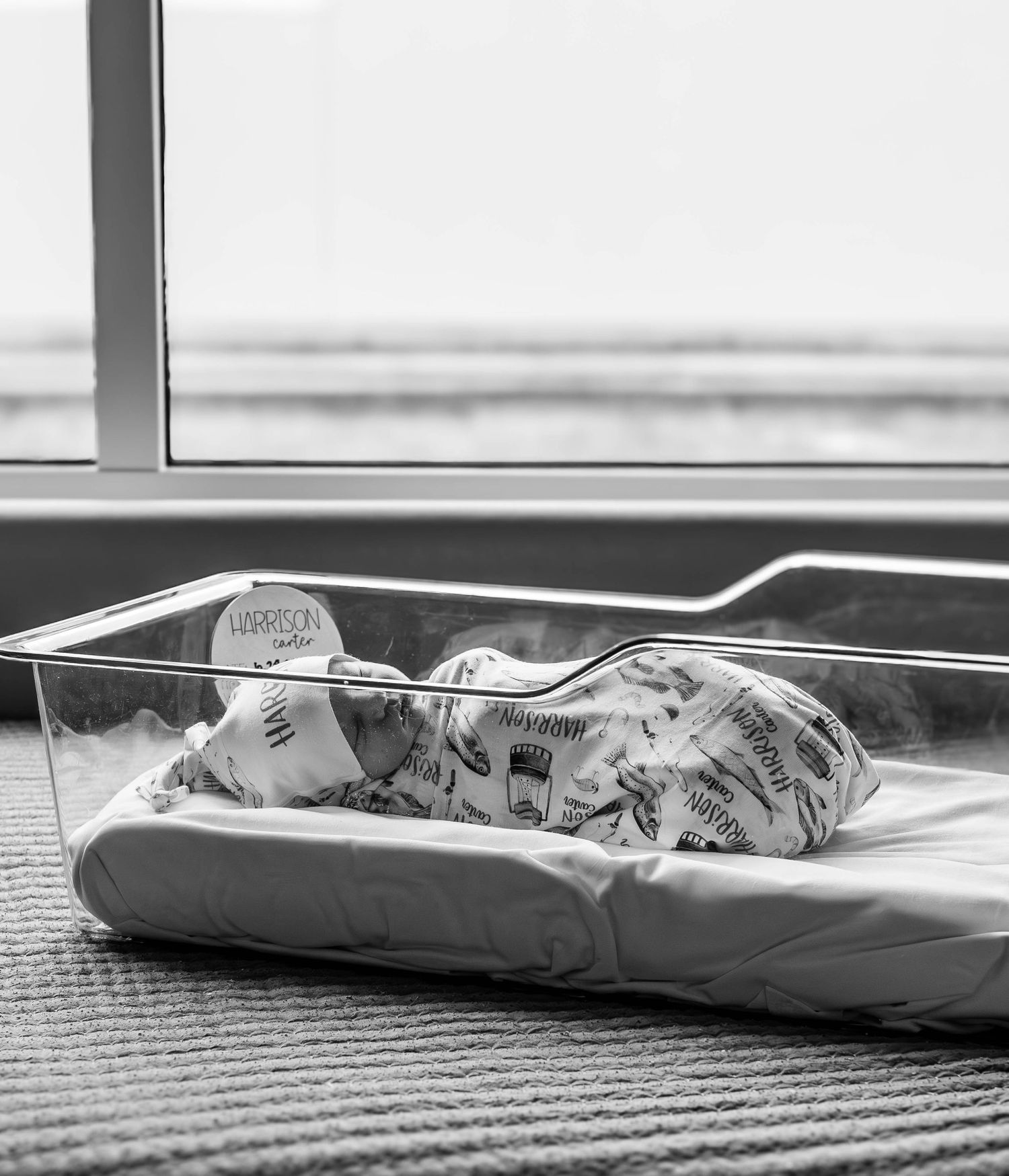 Newborn baby sleeping peacefully in a clear hospital bassinet by a window, wrapped in a patterned swaddle blanket.