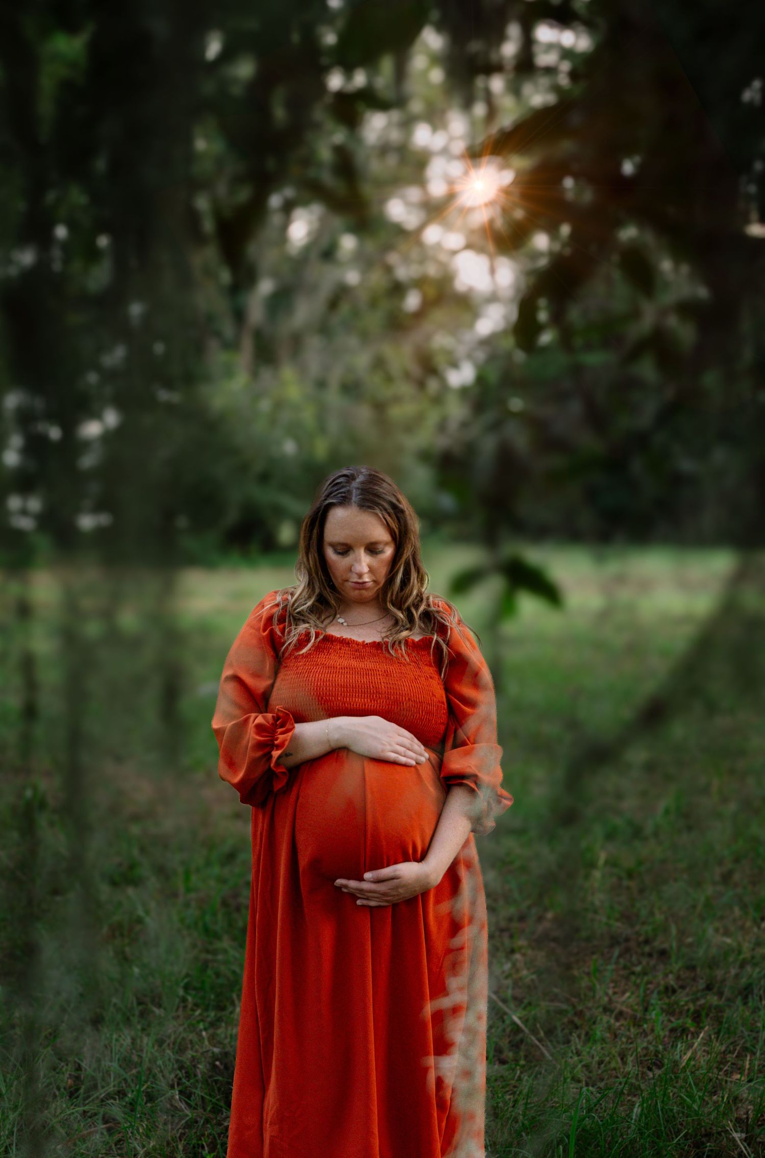 Pregnant woman in red dress cradling baby bump outdoors at sunset in lush green forest setting.