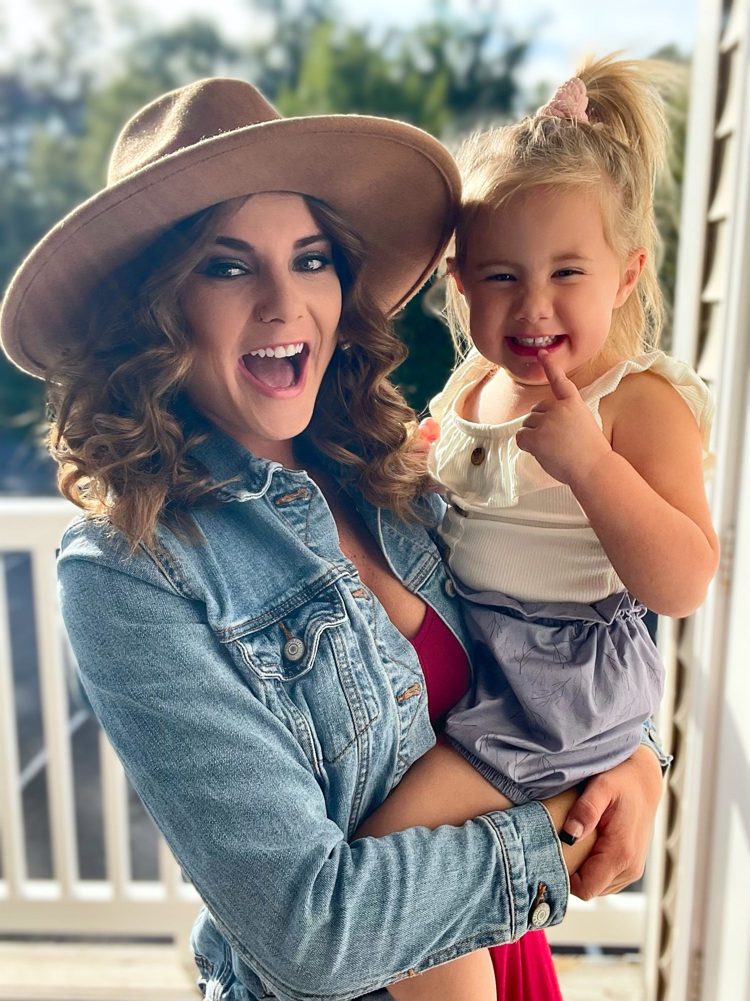 Smiling woman in wide-brim hat holding laughing toddler girl on sunny outdoor porch.