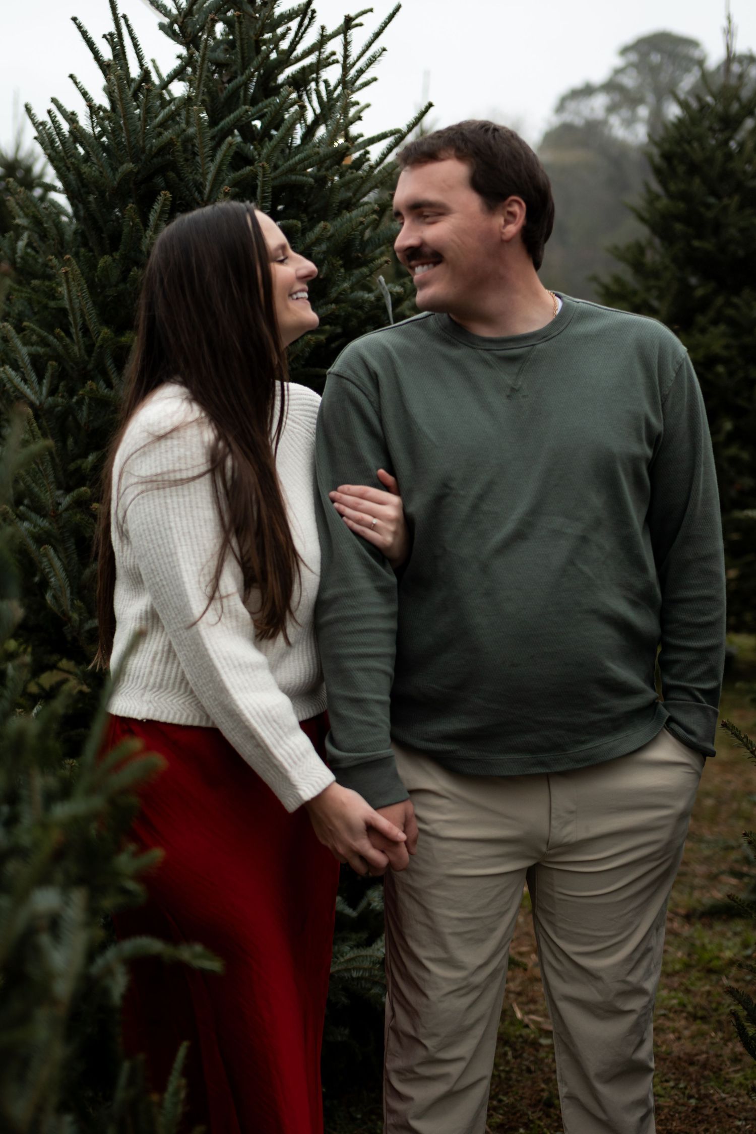 A couple holds hands and smiles at each other among green Christmas trees at a festive tree farm.