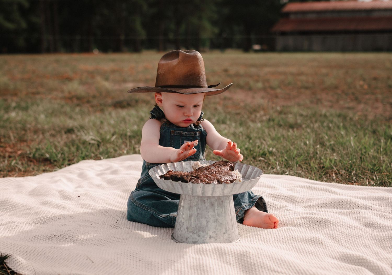 A baby in a cowboy hat and denim overalls sits on a blanket outdoors, eating chocolate smash cake.