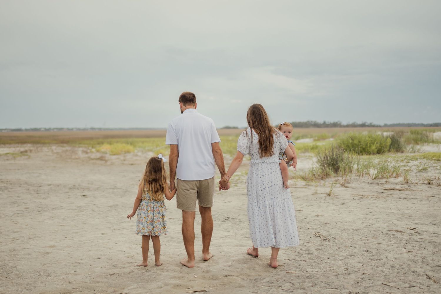 Family of four walking hand-in-hand on a sandy beach, viewed from behind on a cloudy day.