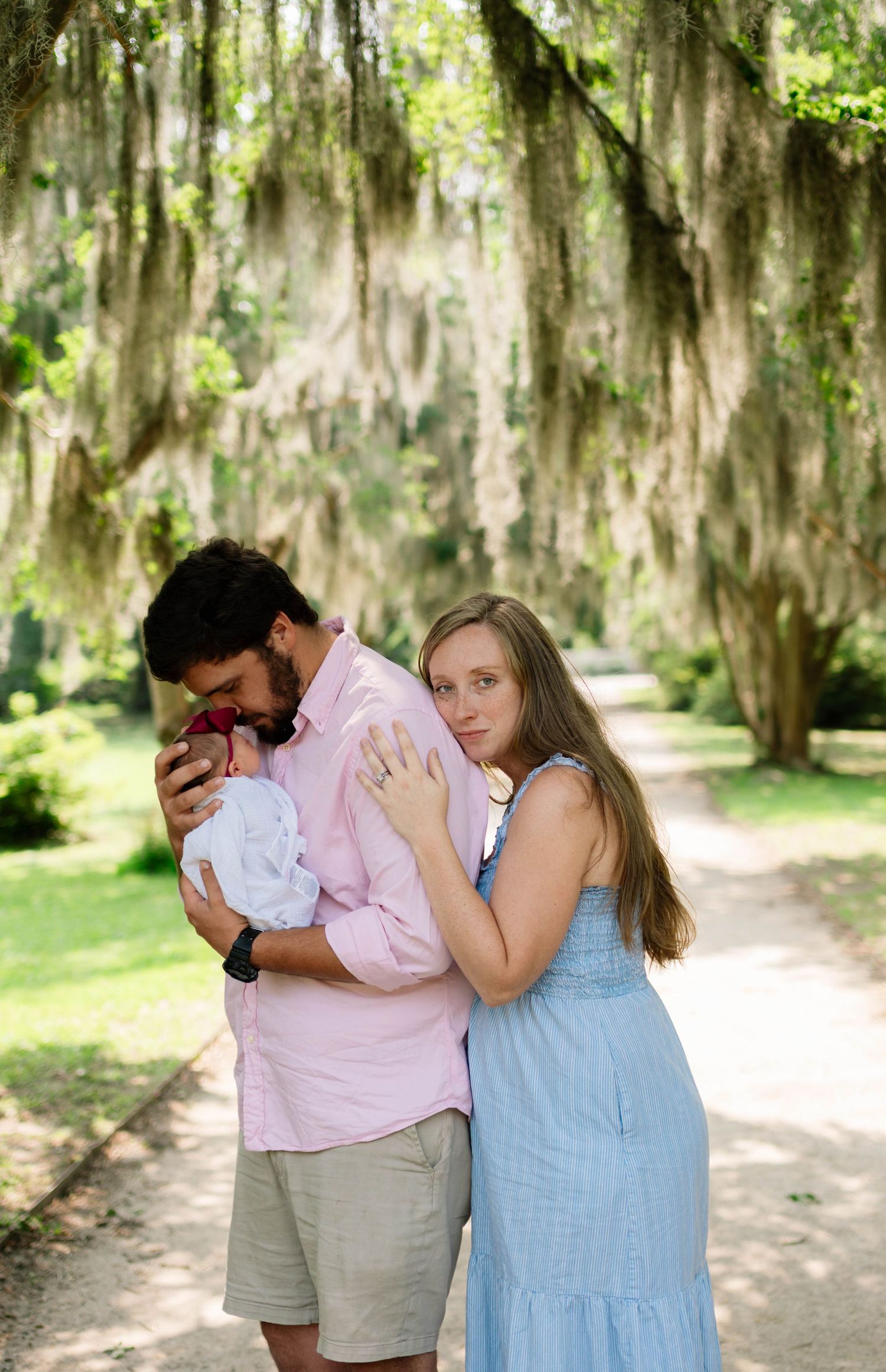 A couple tenderly holds their newborn baby outdoors under Spanish moss-draped trees on a scenic path.