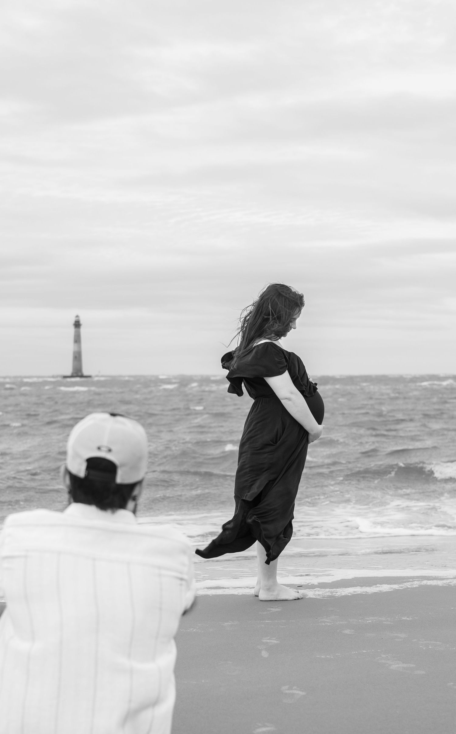 Black and white photo of a woman in a dark dress posing on a beach while a photographer watches, lighthouse in background.