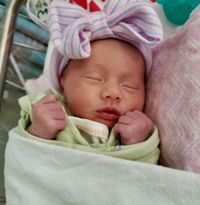 A peaceful newborn baby girl wearing a striped hospital hat sleeps swaddled in a soft blanket.