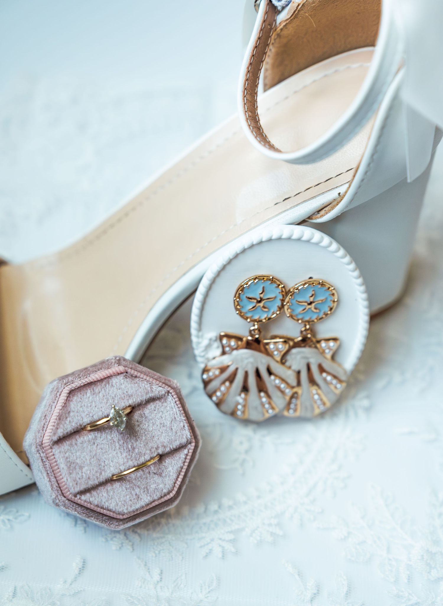 Elegant bridal flat lay with white patent heels, blue and gold star earrings, and a diamond ring in a pink velvet box.