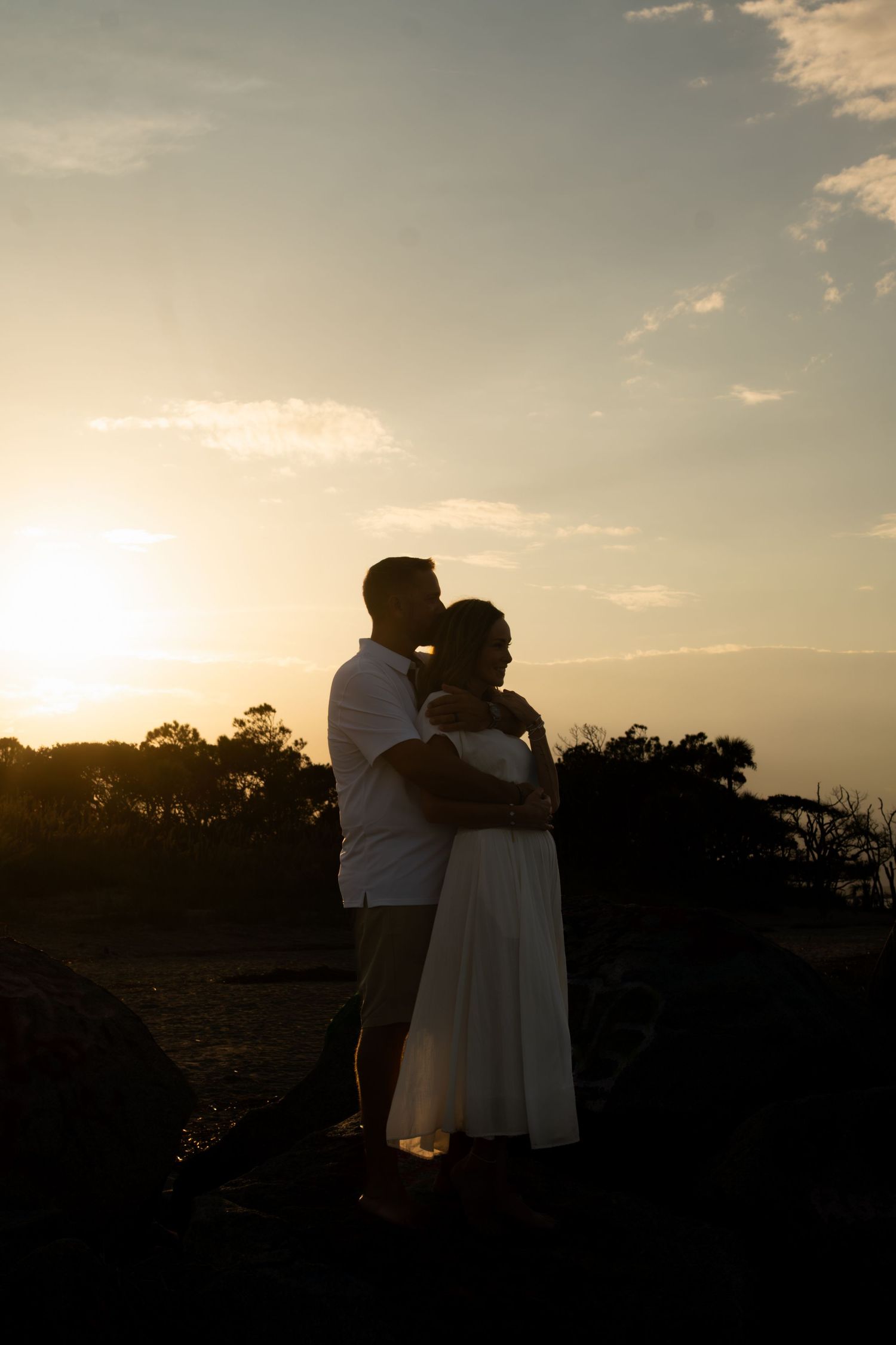 Romantic couple silhouette embracing at golden sunset, man kissing woman in white dress outdoors.