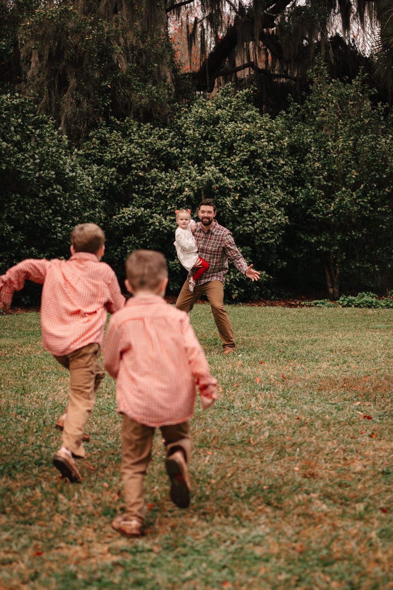 A father joyfully opens his arms to greet his children running toward him on a green lawn.