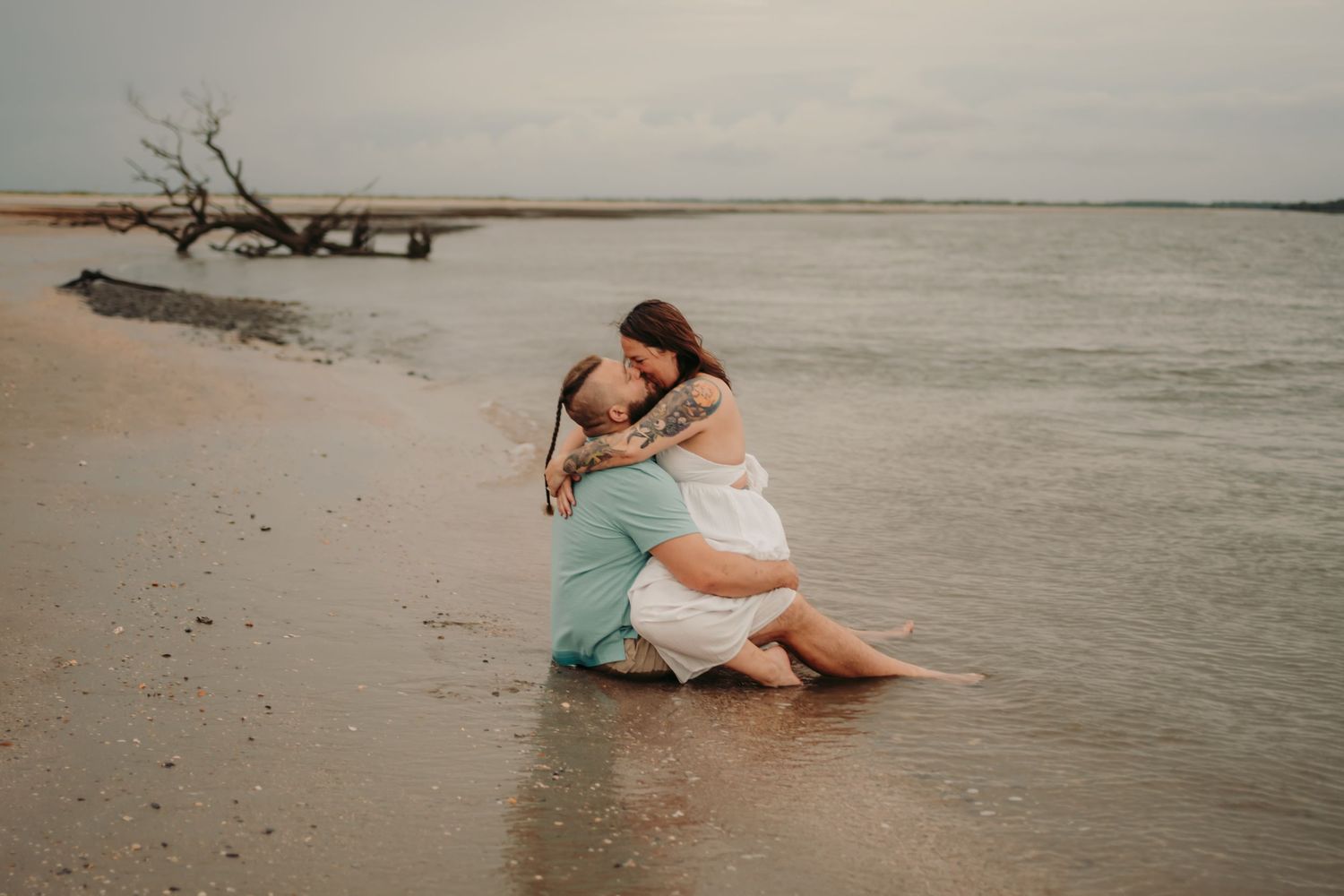 Couple embracing romantically on a sandy beach shore with calm ocean waters and driftwood in the background.