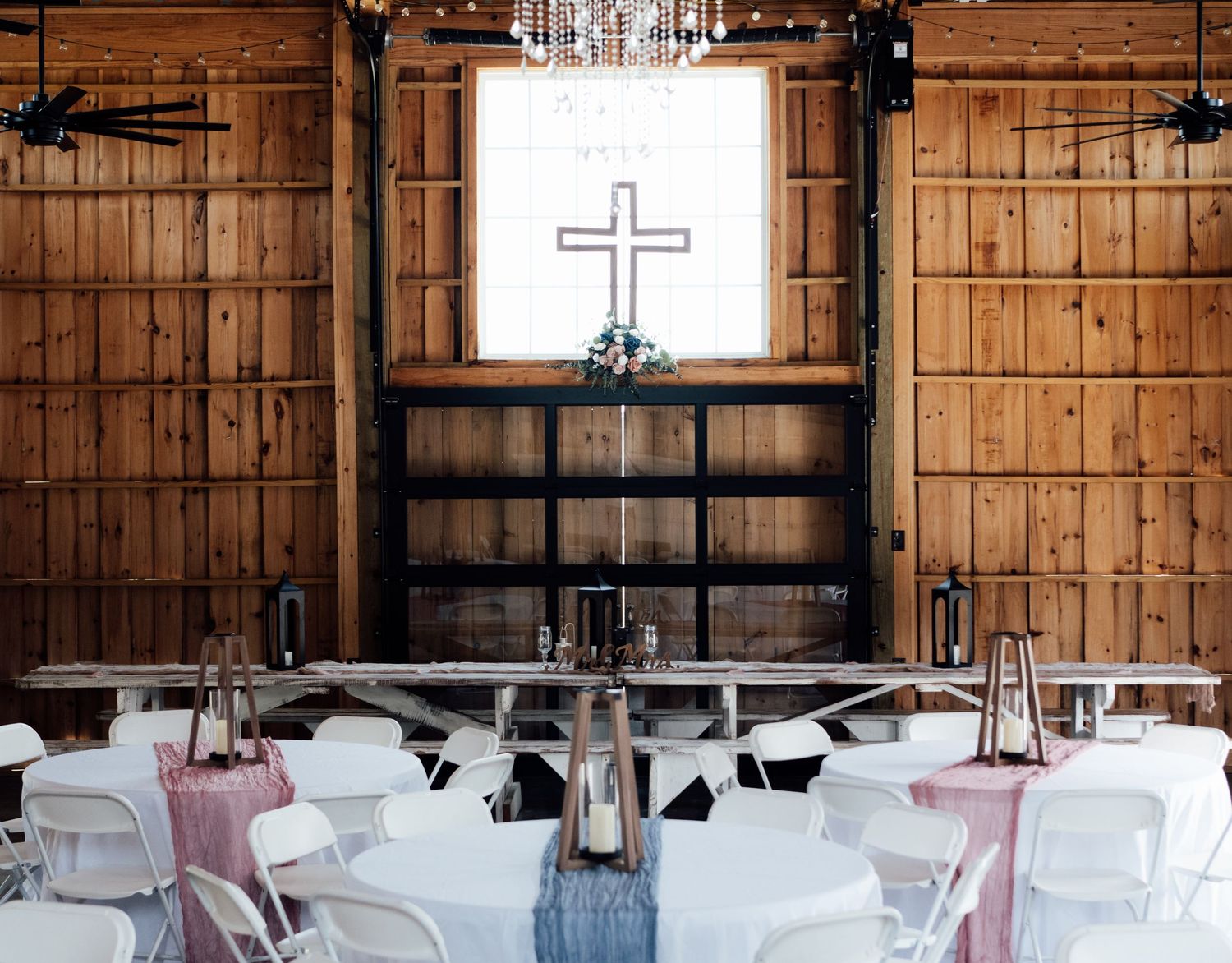 Rustic barn wedding reception with white chairs, wooden tables, cross backdrop, string lights, and pink table runners.