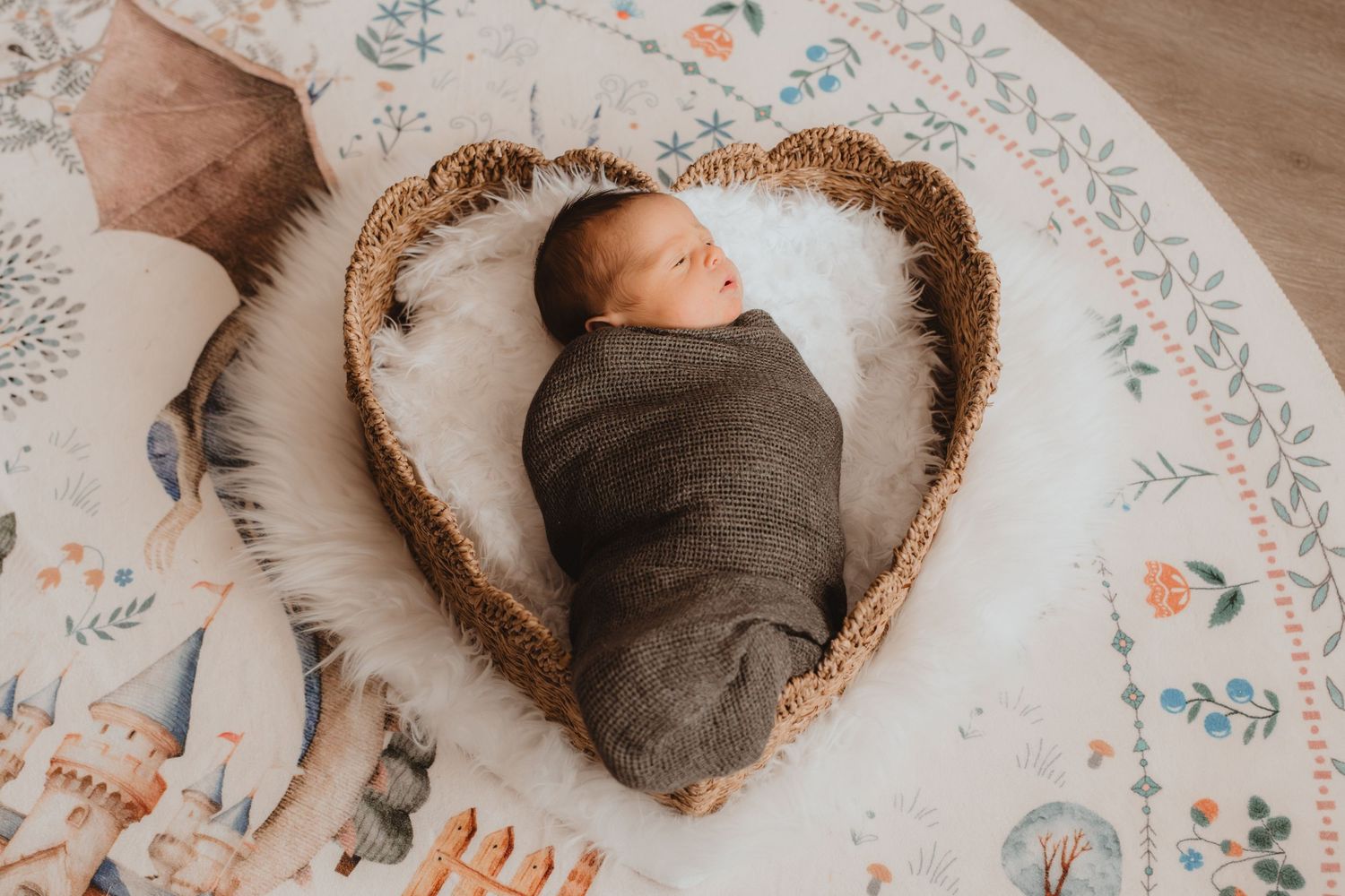 Newborn baby swaddled in gray wrap sleeping in heart-shaped wicker basket on floral play mat.
