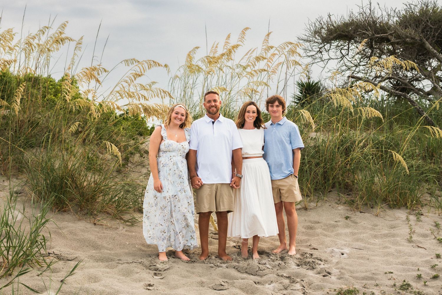 Family of four smiling together on sandy beach dunes with sea oats and greenery in background.