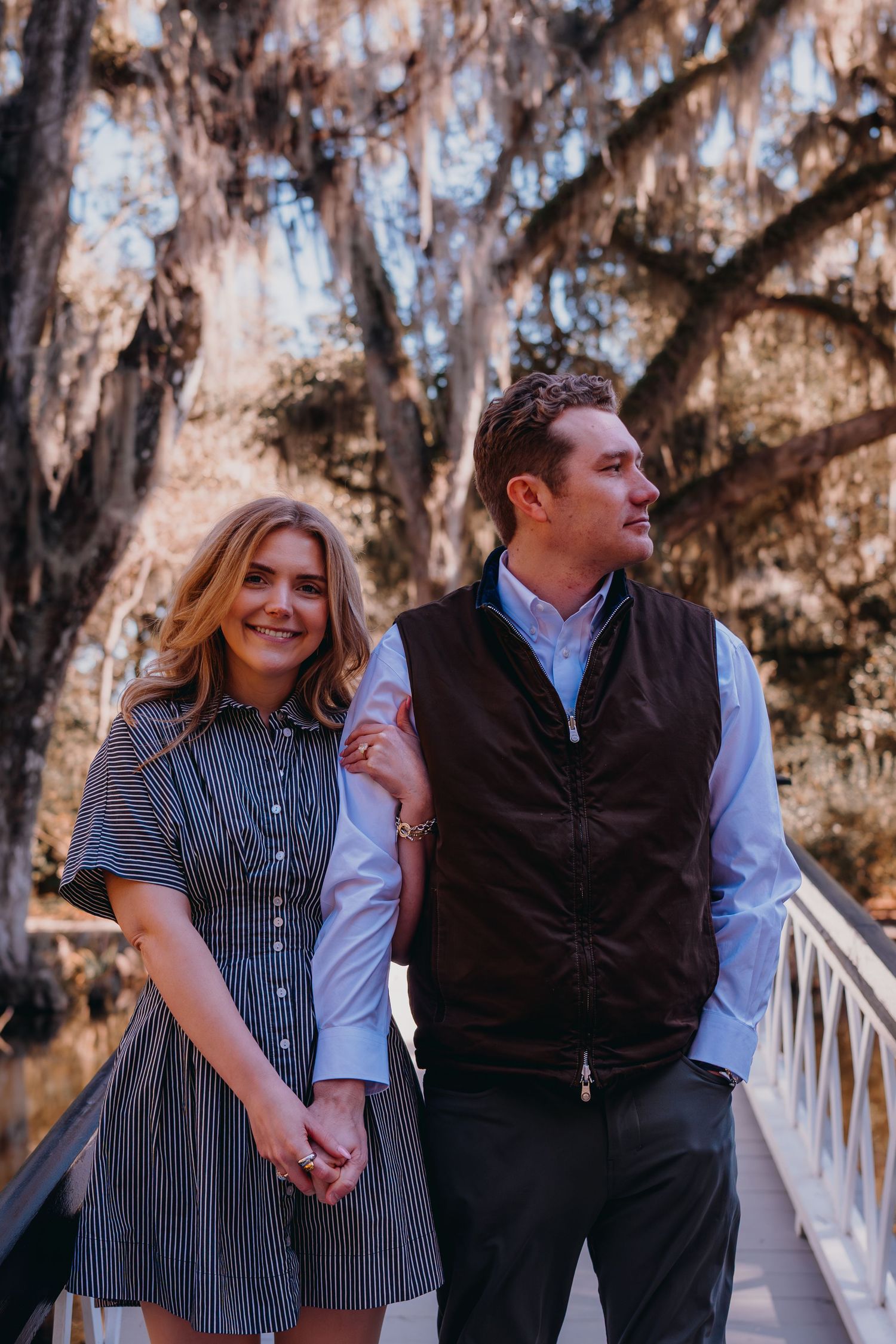 A couple holds hands on a wooden bridge surrounded by Spanish moss-draped trees in a romantic outdoor setting.