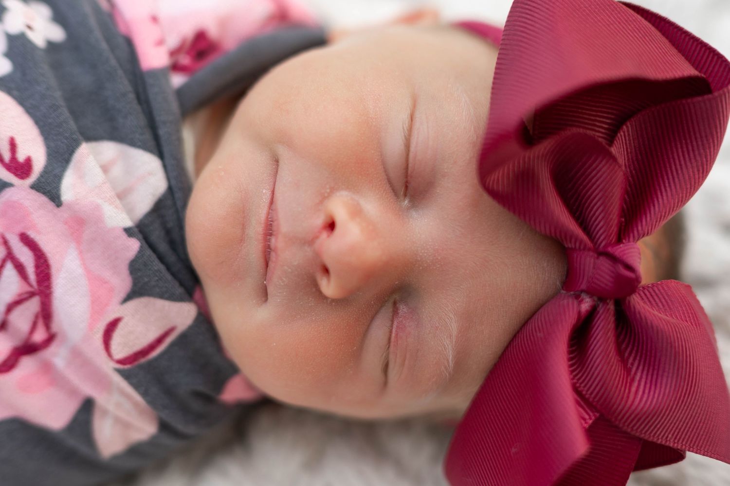 Sleeping newborn baby girl wearing a floral hat and large burgundy bow headband.