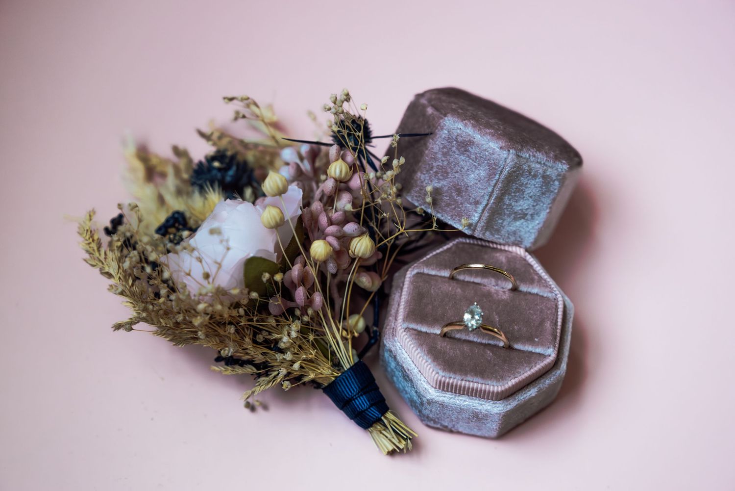 Elegant engagement ring in velvet box beside a dried floral boutonniere on a soft pink background.