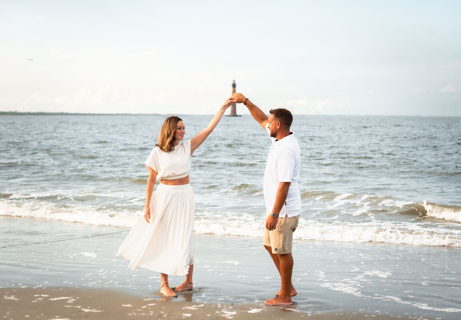 Couple in white outfits dancing on a sandy beach, holding hands raised, with ocean waves in the background.