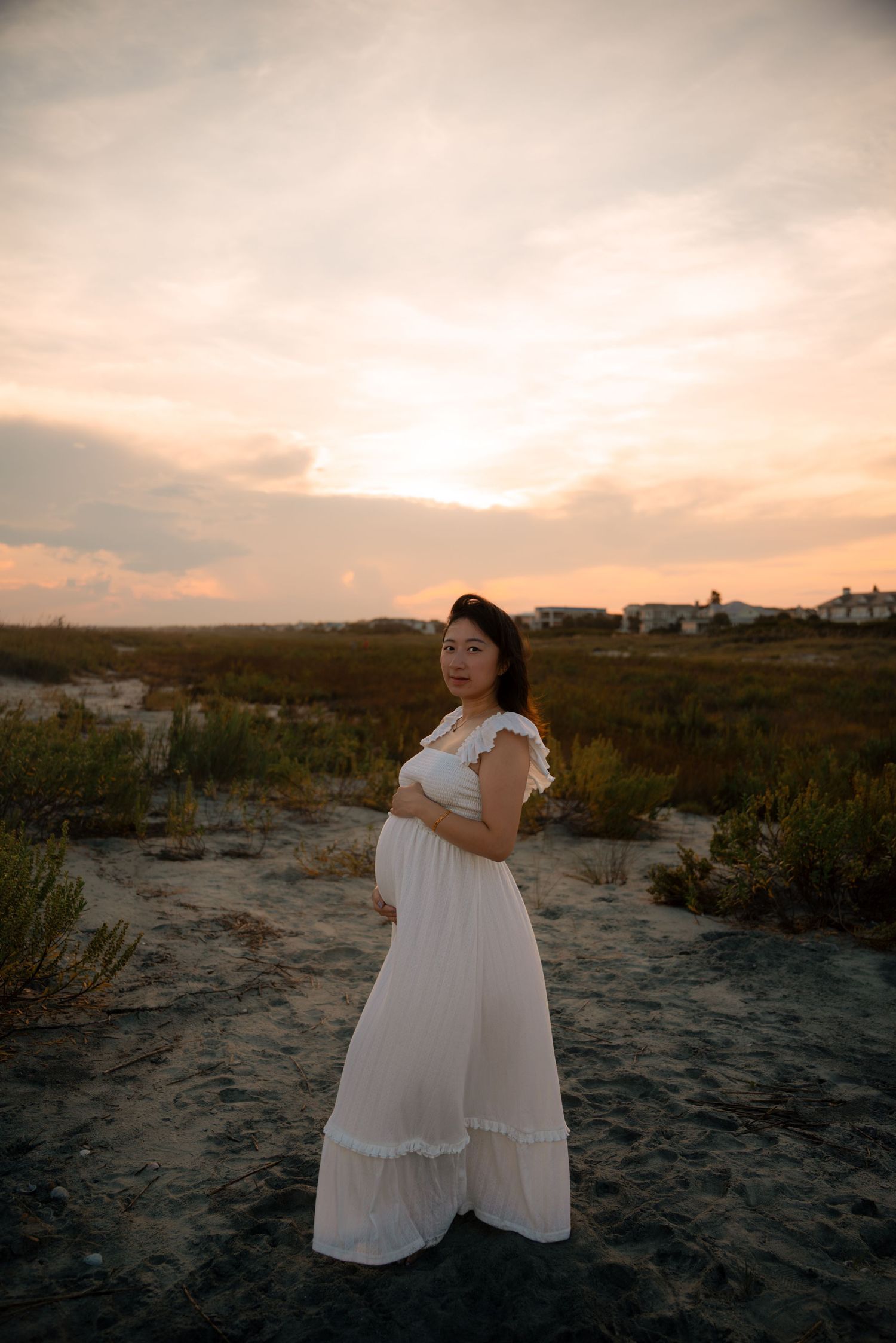 Pregnant woman in white maxi dress poses on sandy beach at sunset with golden sky backdrop.