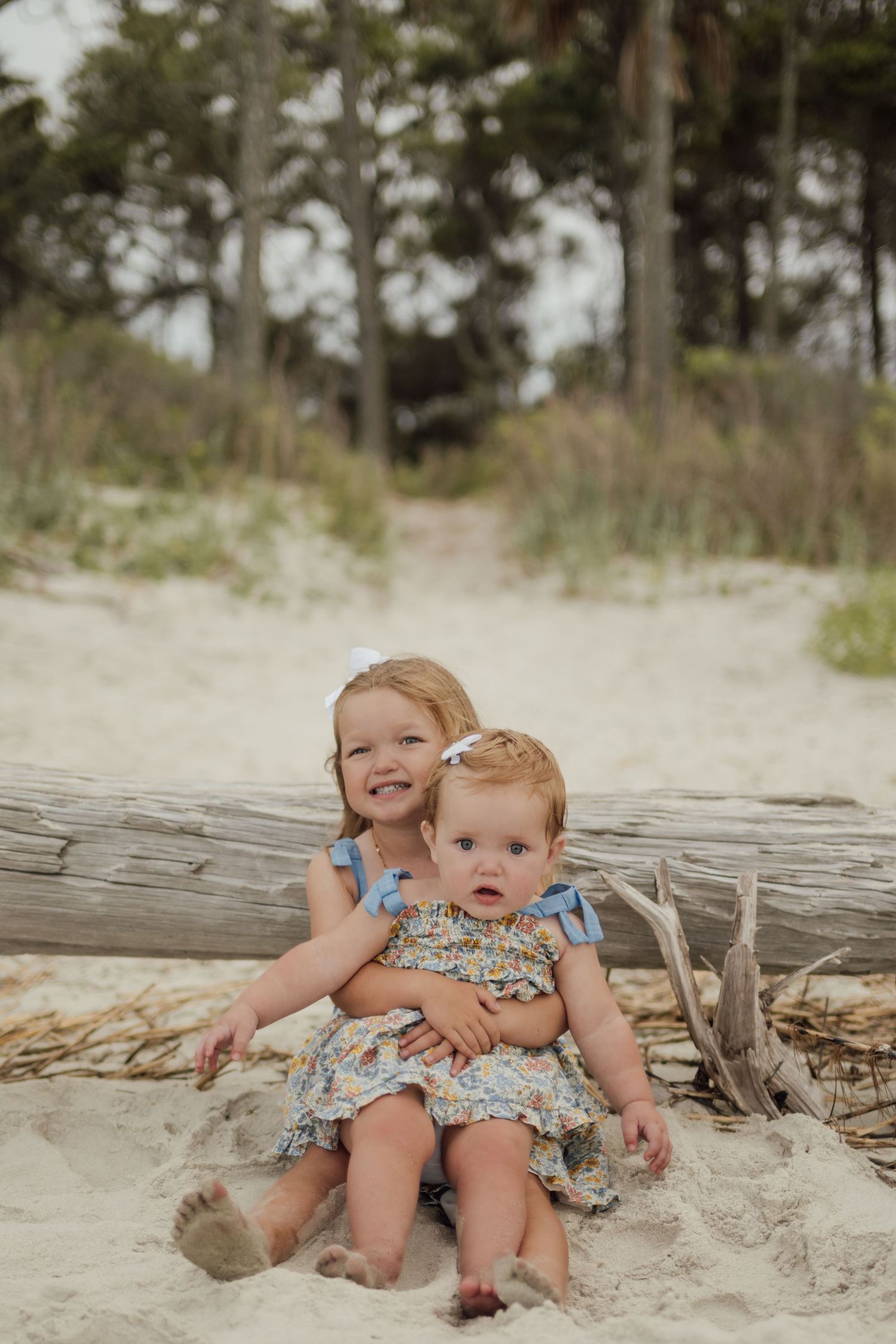 Two young sisters in floral dresses sit together on sandy beach beside driftwood, smiling with trees in background.