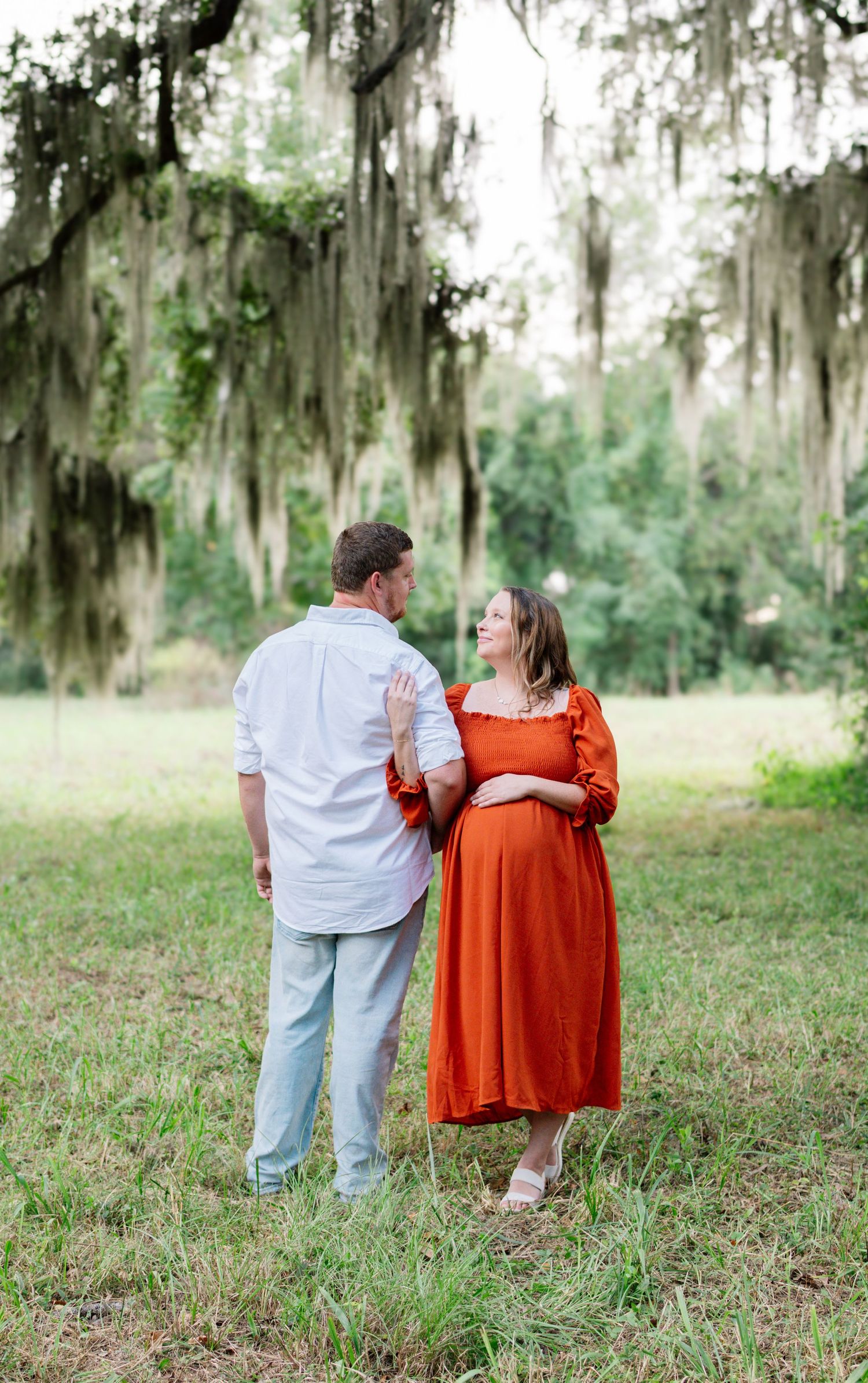 Expecting couple sharing a tender moment in orange dress and blue shirt under Spanish moss trees.