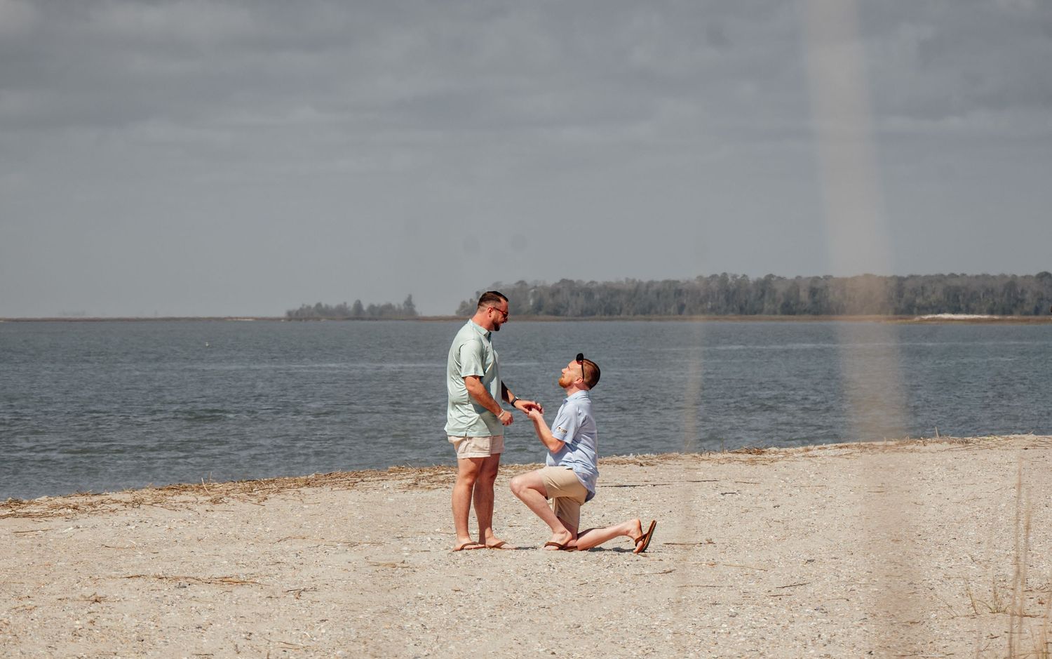 Same-sex couple sharing a romantic beach proposal moment with a rainbow in the background on a sunny day.