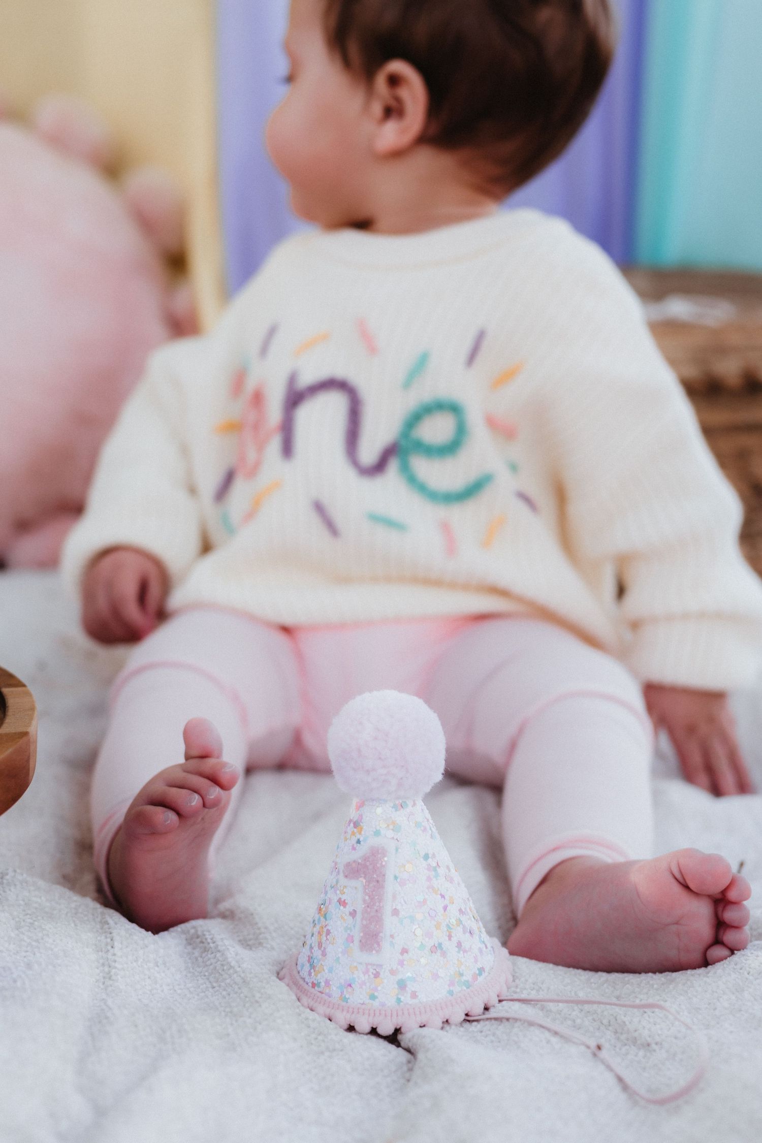Baby in 'one' sweater sits beside a number 1 party hat, celebrating their first birthday milestone.