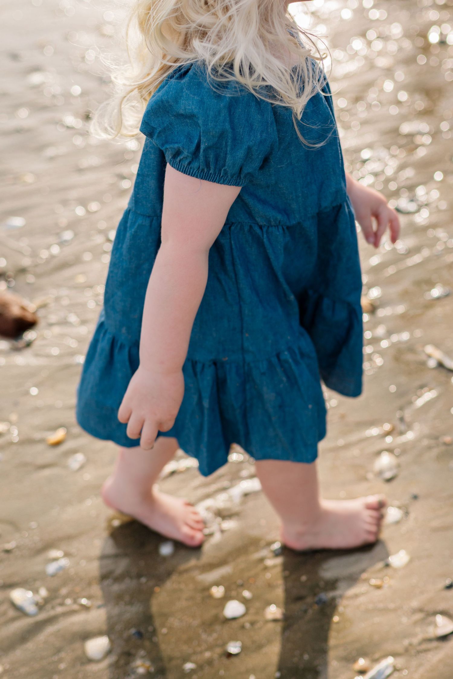Young blonde toddler girl in teal dress walking barefoot on sandy beach with shells.