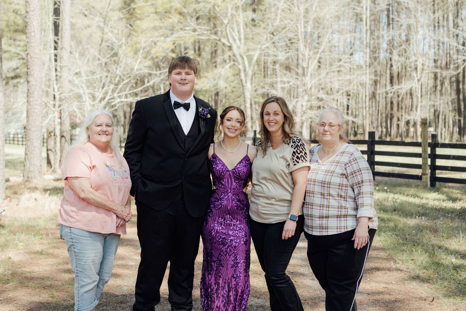Family group of five posing outdoors in formal attire near a wooden fence with bare trees in background.