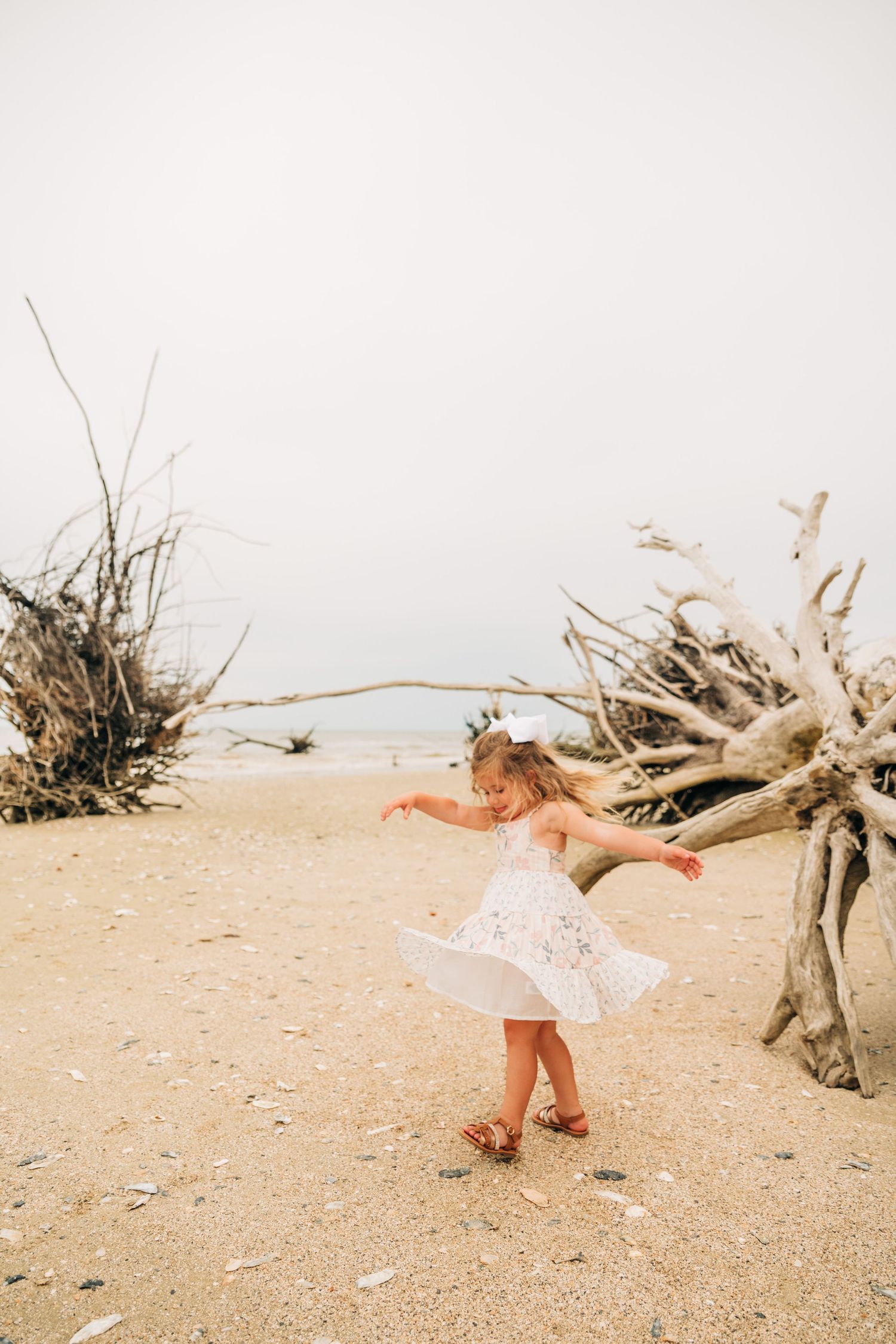 A young girl in a white dress and bow spins joyfully on a sandy beach near weathered driftwood.
