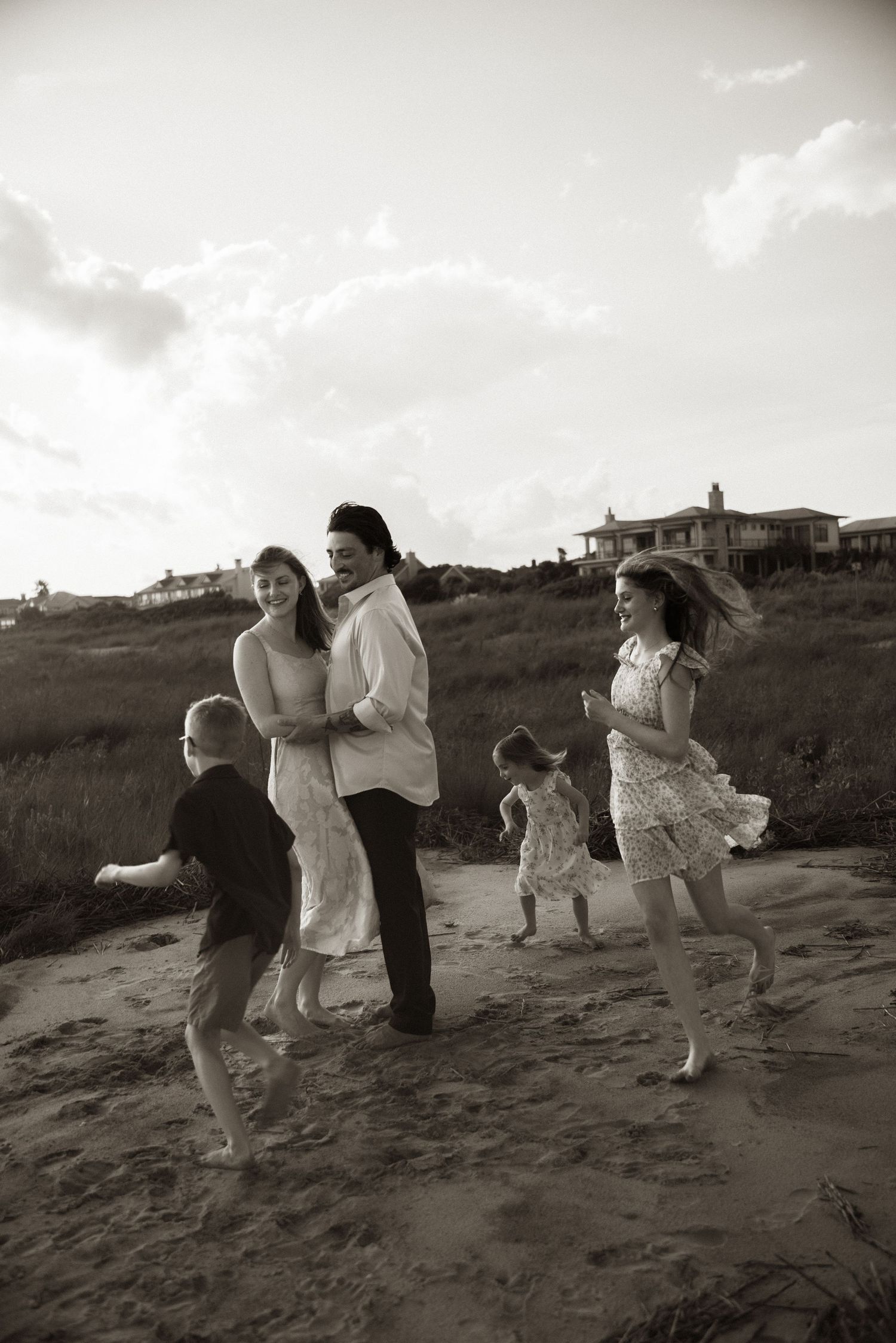 Black and white photo of a joyful family playing on a sandy beach path with children running and parents embracing.