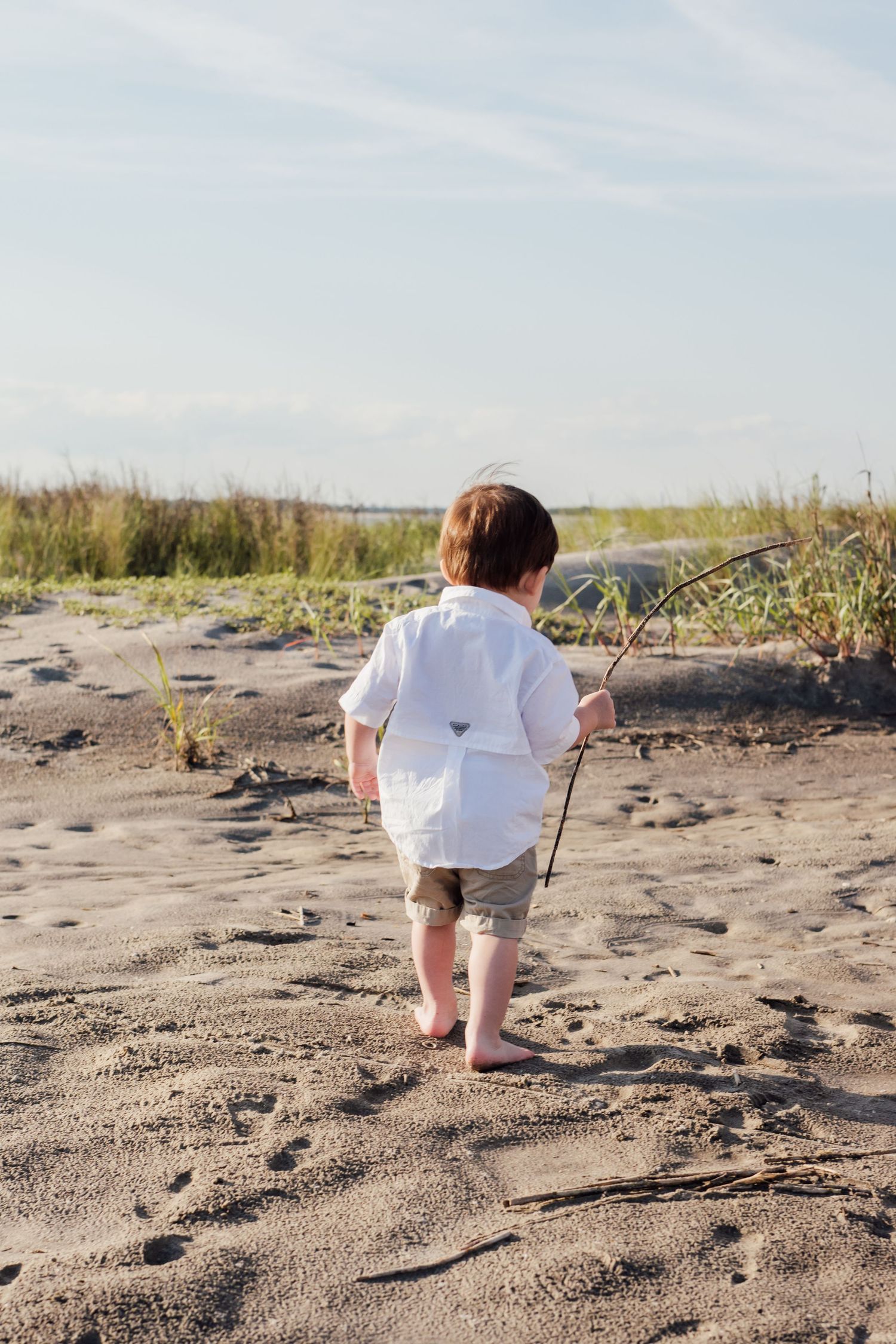 Young boy in white shirt walks barefoot on sandy beach holding a stick, viewed from behind.