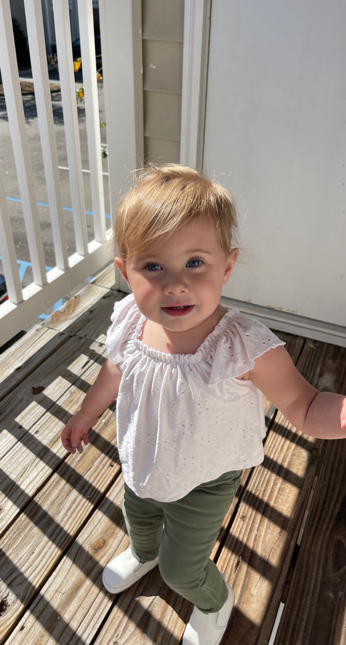 Sweet blonde toddler in white top and green pants standing on sunny wooden deck.