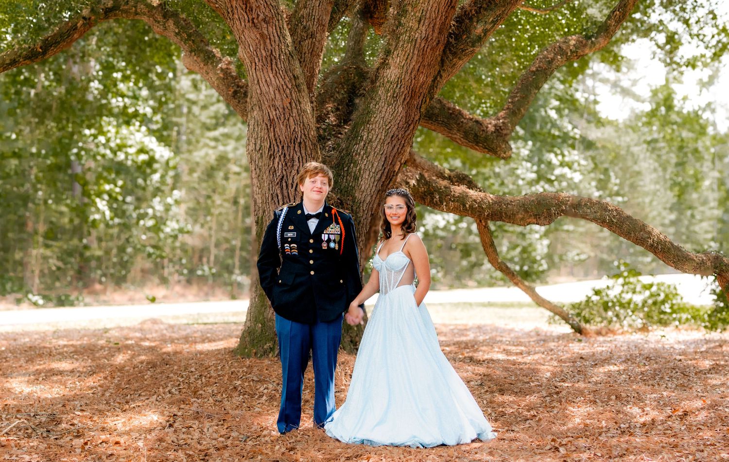 A couple in formal attire, military uniform and blue gown, pose under a large oak tree outdoors.