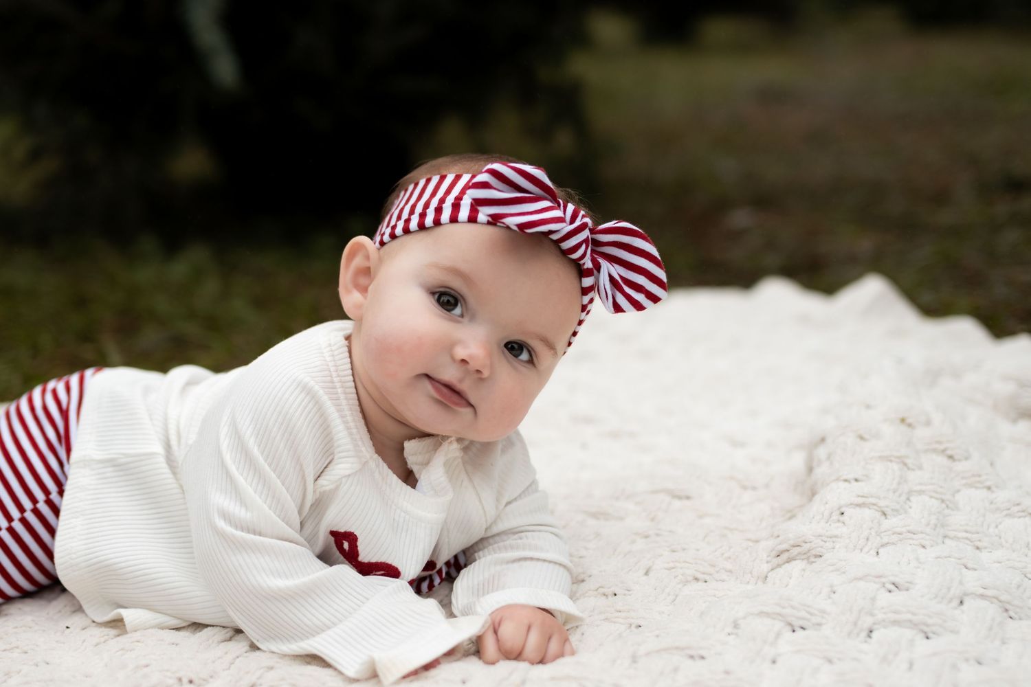 A cute baby in a white outfit and red striped bow headband doing tummy time on a white blanket outdoors.