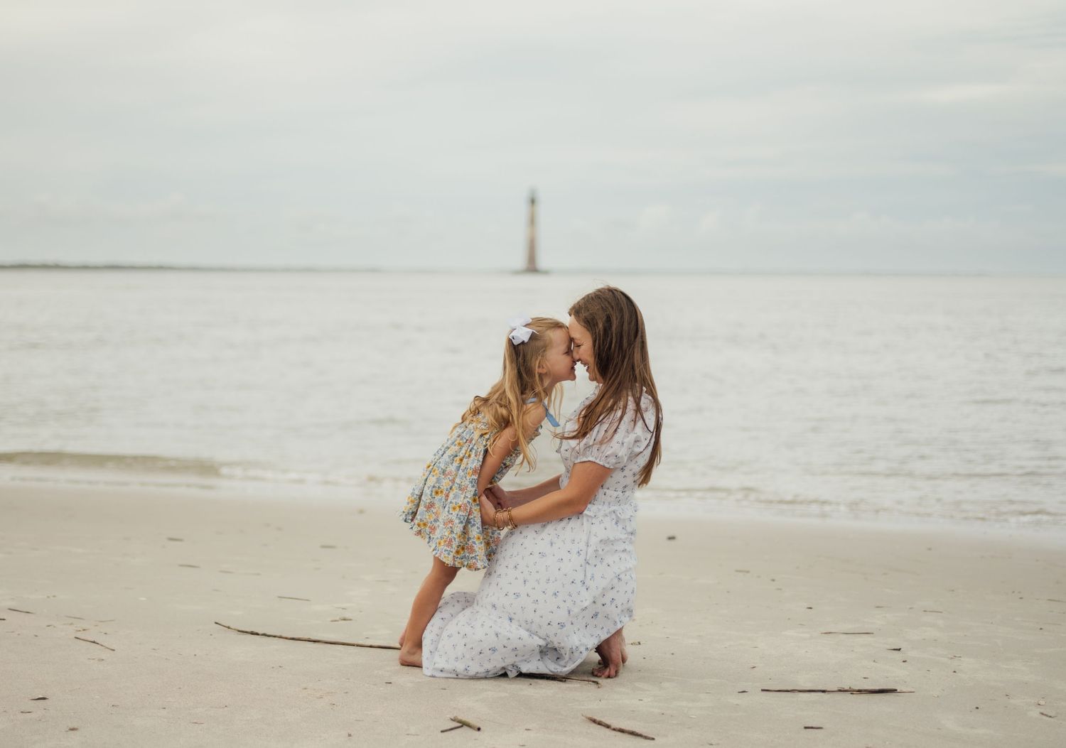 Mother and young daughter share a tender kiss on a sandy beach with a lighthouse in the background.