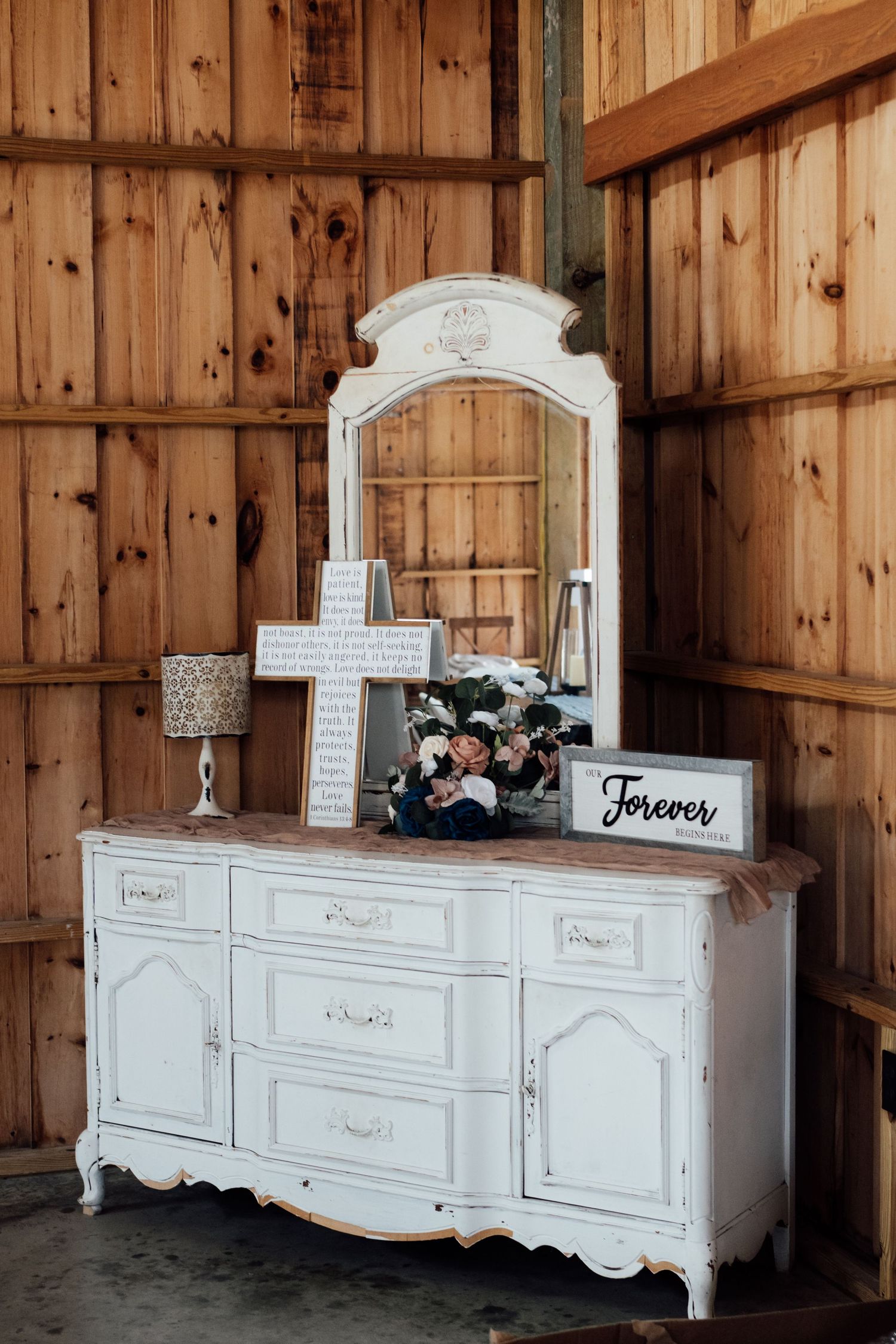 White vintage dresser with mirror displaying wedding decor, floral arrangement, and Forever sign in rustic barn setting.