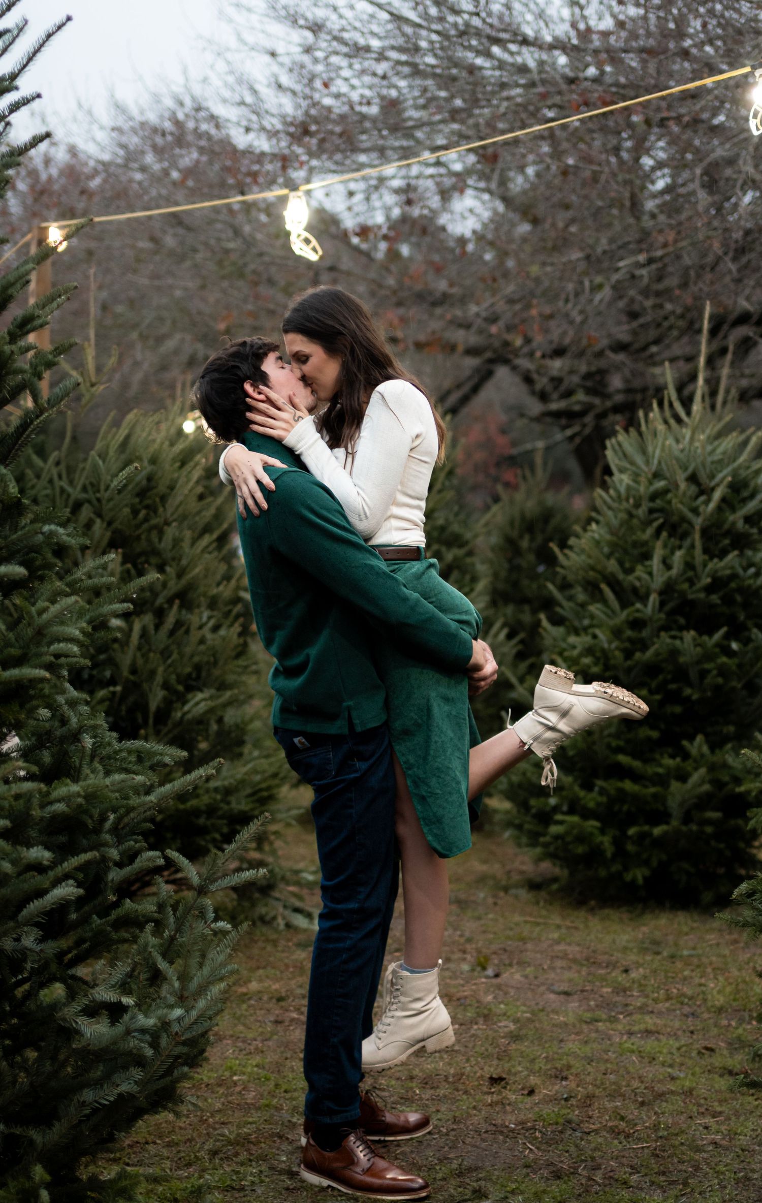 Couple sharing a romantic kiss at a Christmas tree farm, surrounded by evergreens and string lights.