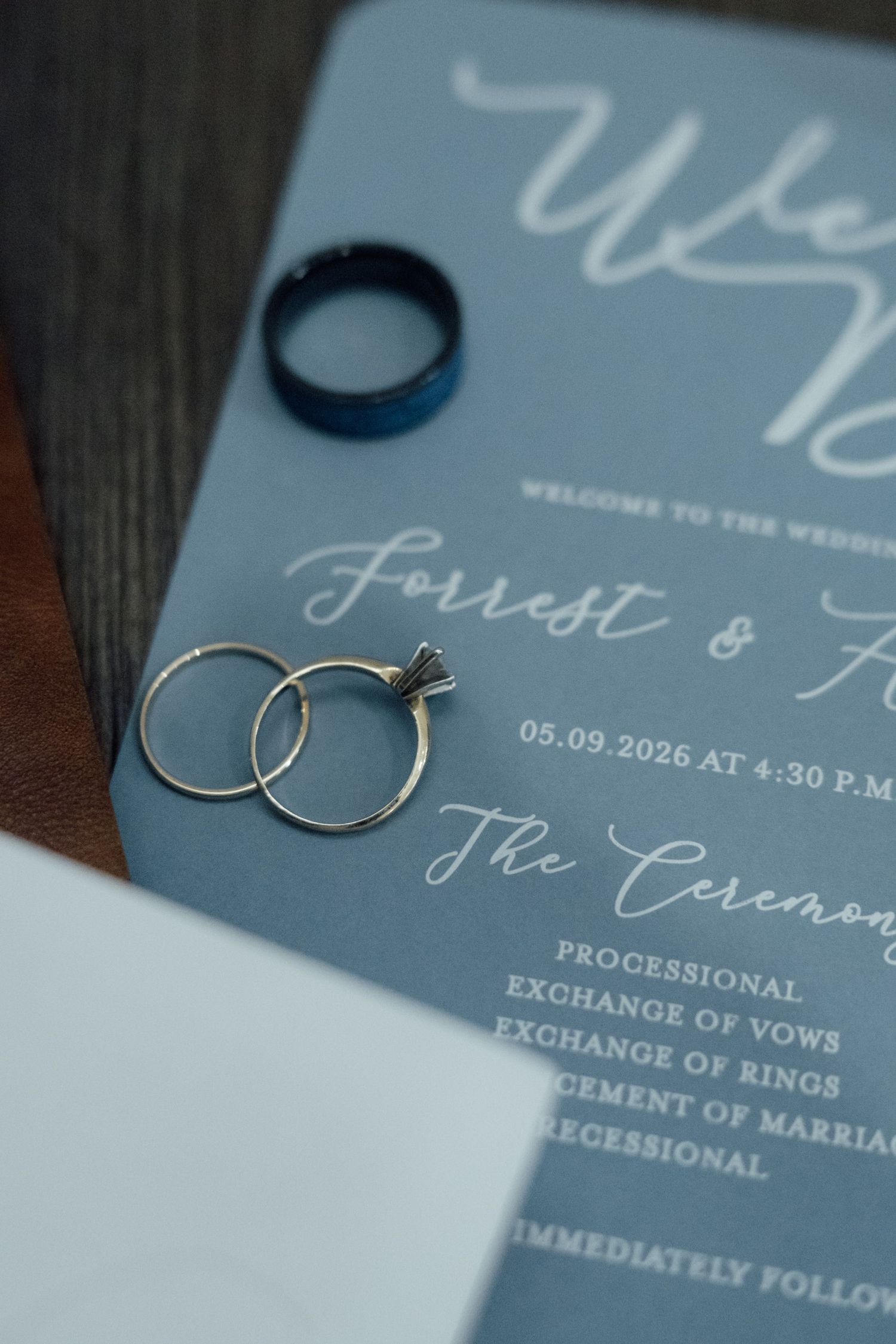 Dusty blue wedding program with gold engagement rings and blue band, featuring Forrest and Ashley ceremony details dated 05.09.2026.