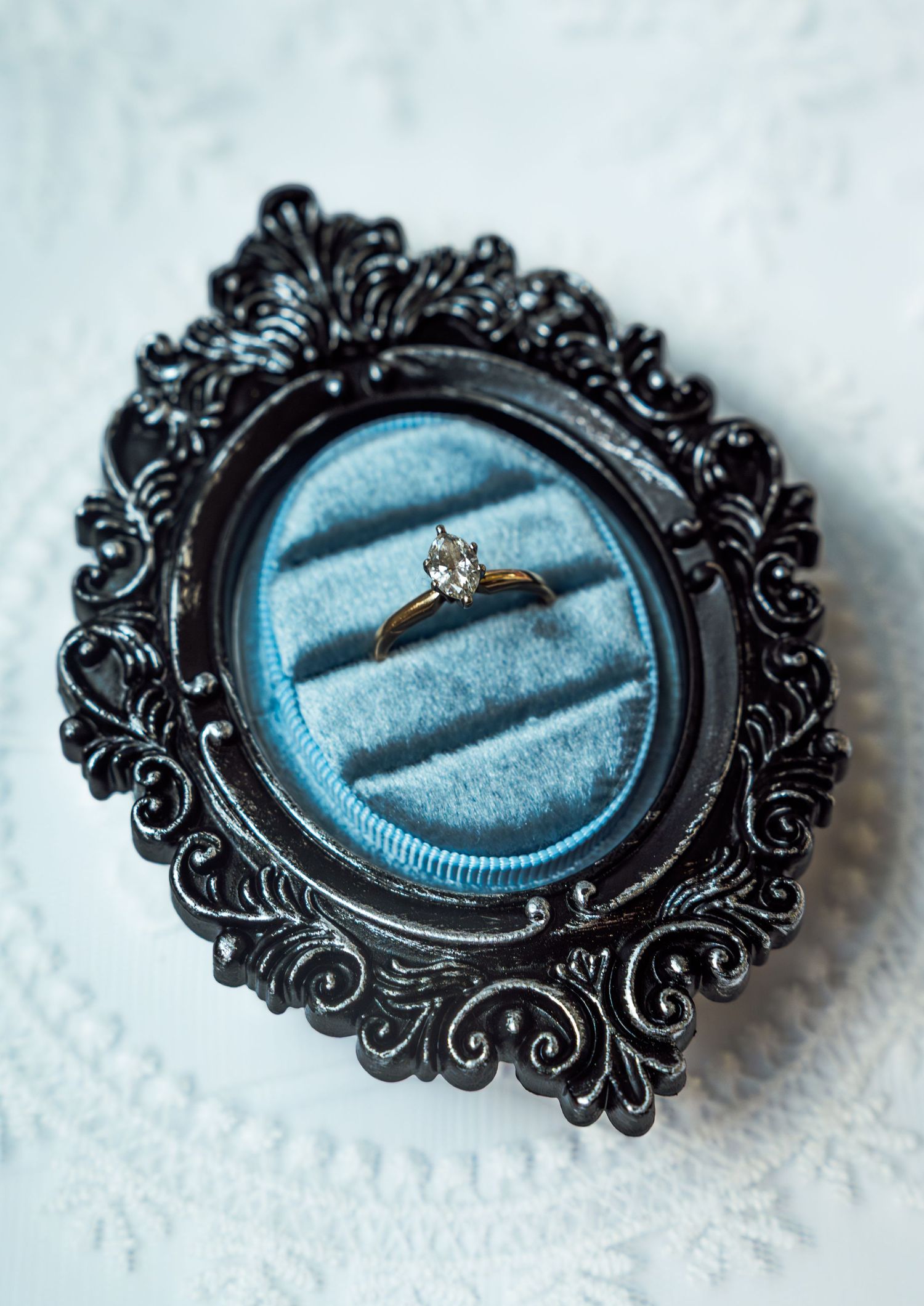 Gold solitaire diamond engagement ring displayed in ornate black baroque ring box with blue velvet interior.