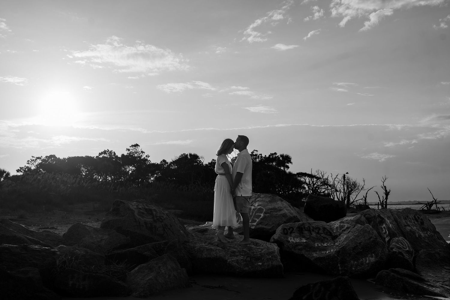 Black and white photo of a couple sharing a romantic moment on rocks at sunset near the ocean.