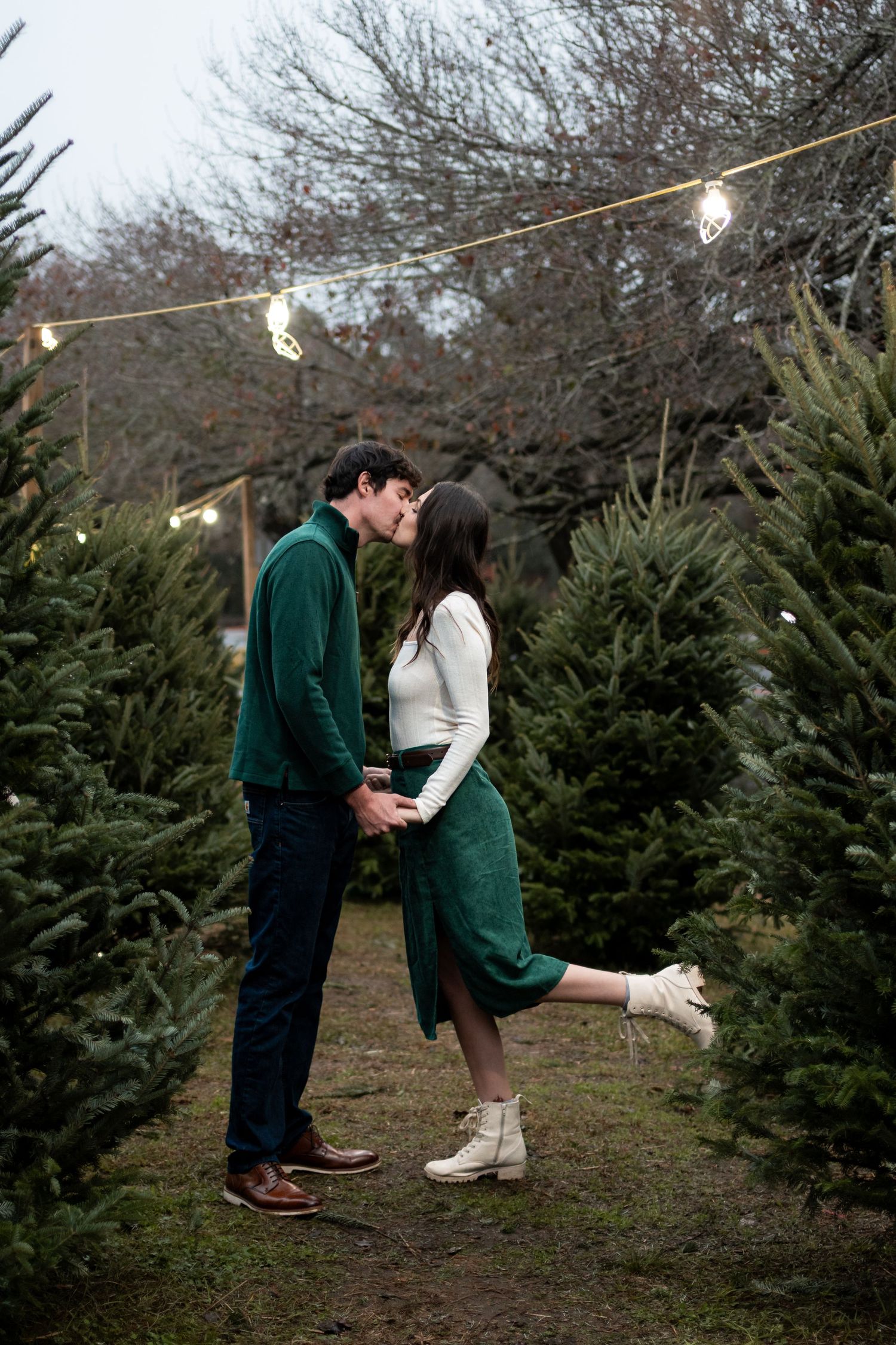 Couple sharing a romantic kiss at a Christmas tree farm, surrounded by green trees and string lights.