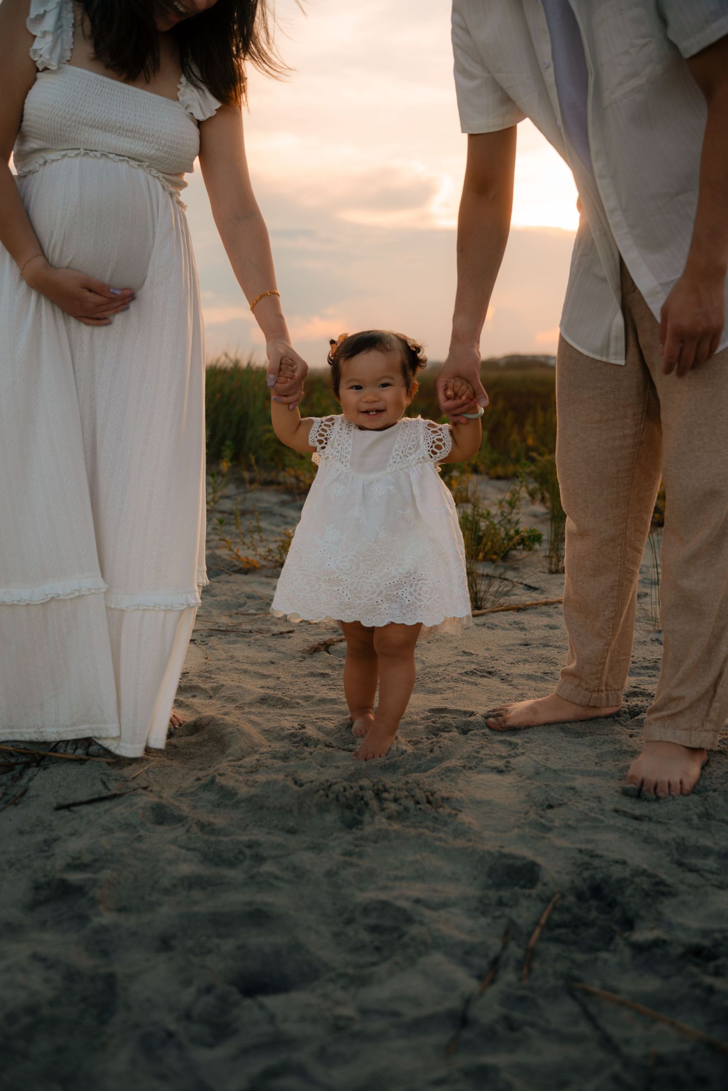 A toddler in a white dress holds her parents hands on a sandy beach at sunset, with her pregnant mother beside her.