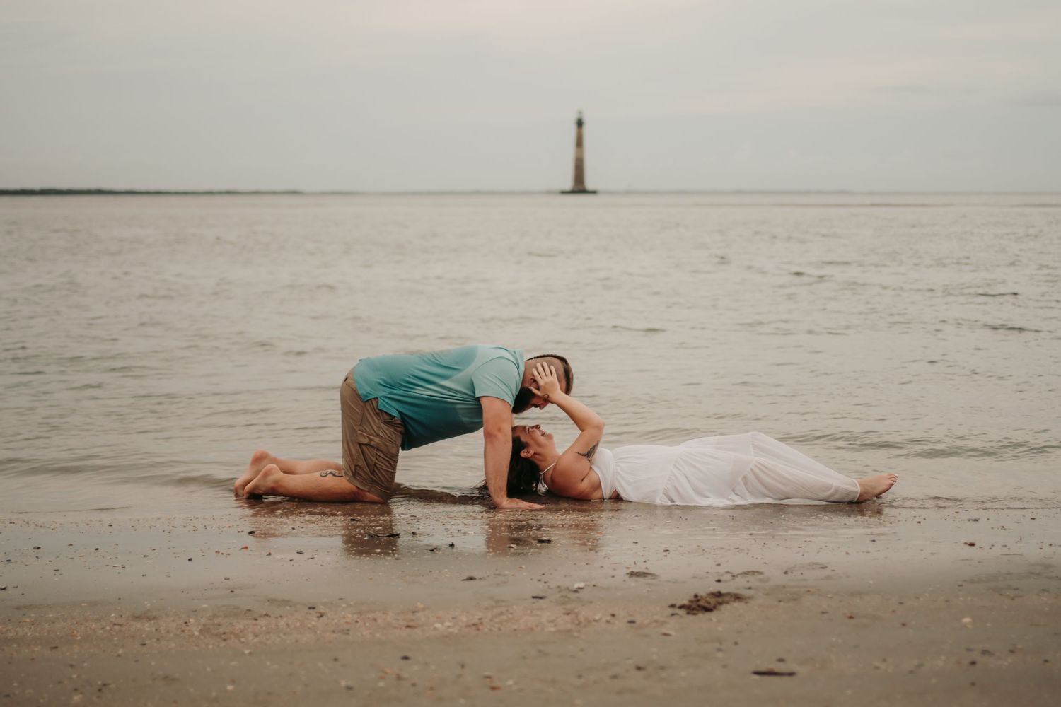 Couple sharing romantic moment on beach shore with lighthouse in background on overcast day.