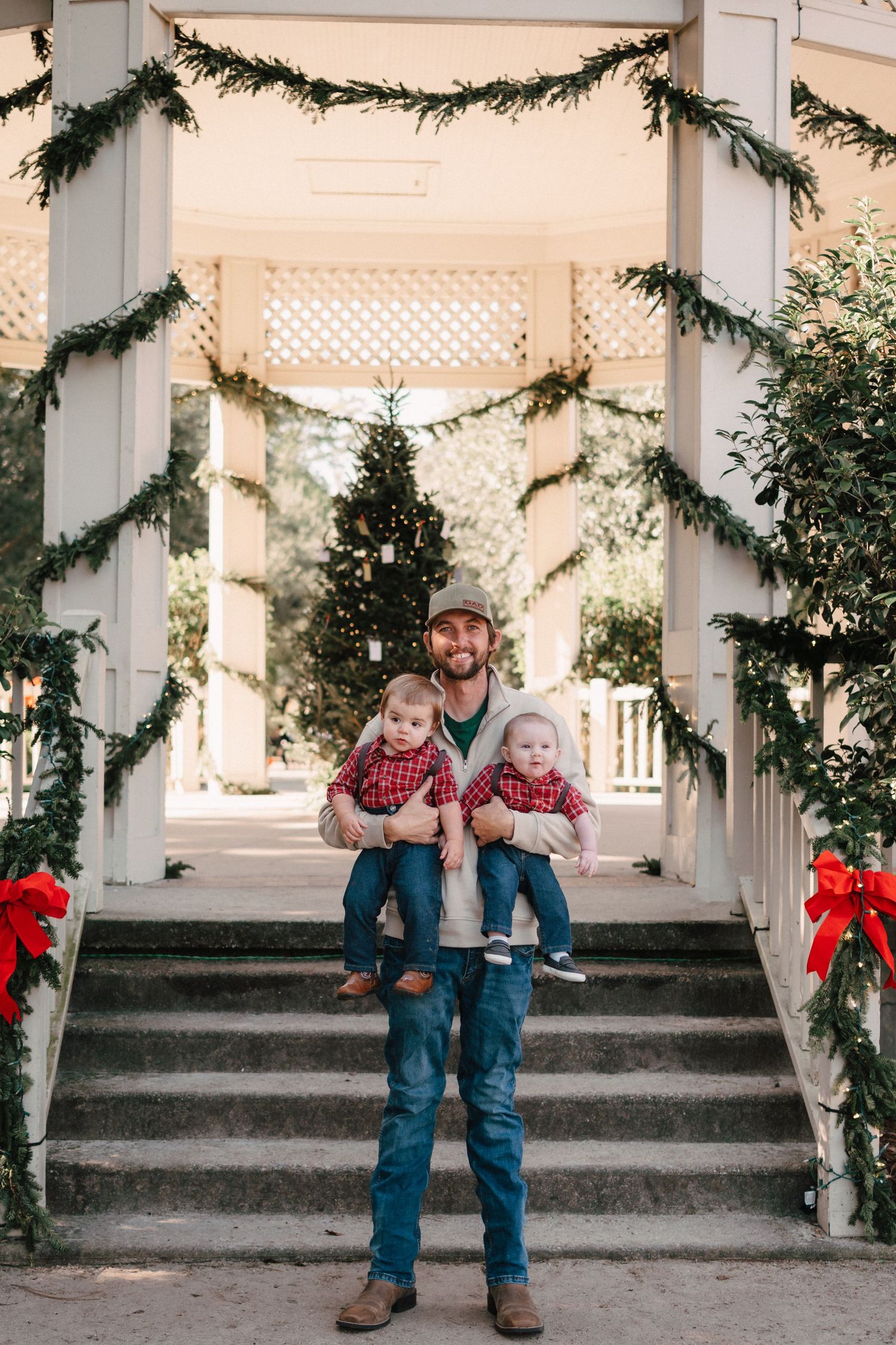 Father holding two toddlers in matching red plaid outfits on Christmas decorated steps with garland and bows.
