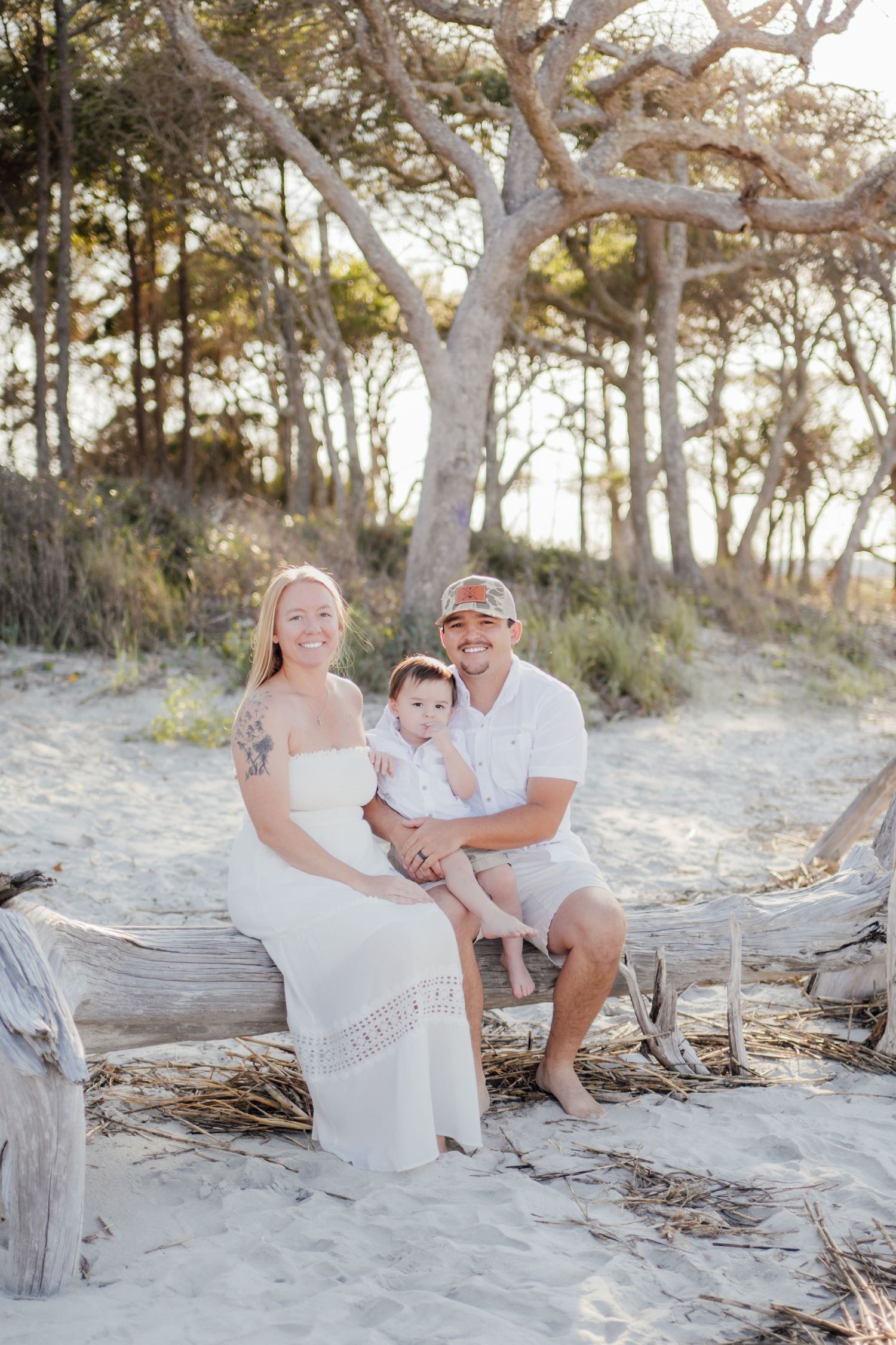 A smiling family of three poses on driftwood at a sandy beach, with a baby in white and trees in the background.